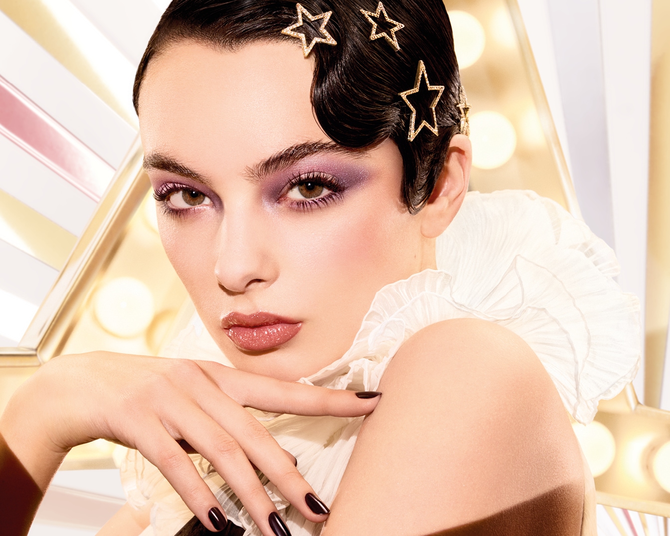 Primo piano di una modella con ombretto viola, labbra lucide e fermagli a forma di stella dorata tra i capelli, che mette in risalto un look glamour e sofisticato.