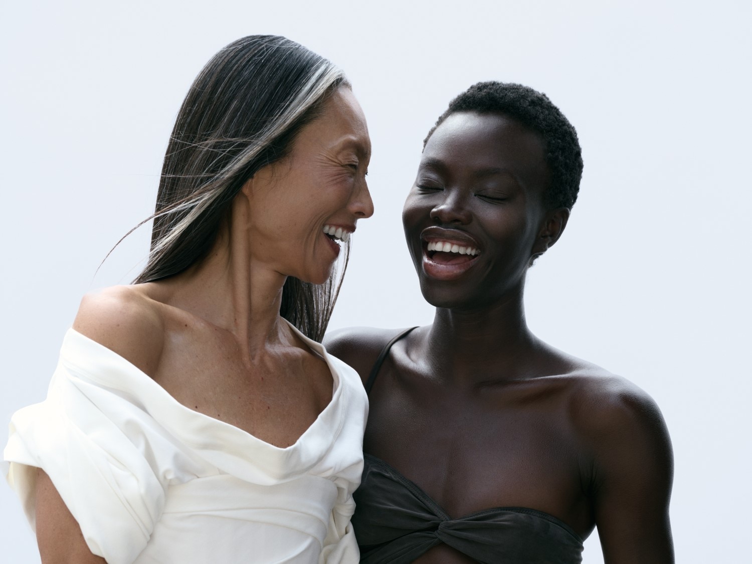 Zwei lachende Frauen unterschiedlicher Hautfarbe, die eine mit grau meliertem, langem Haar und weißem Oberteil, die andere mit kurzem, dunklem Haar und dunklem Top.