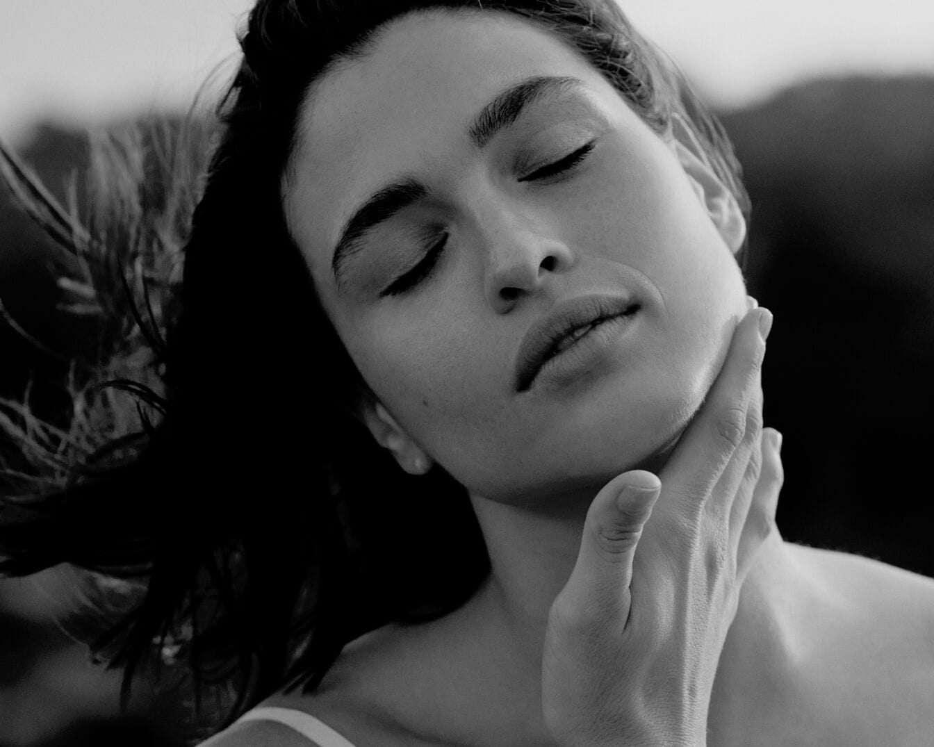 Na czarno-białym zdjęciu widać kobietę z zamkniętymi oczami, która delikatnie dotyka dłonią swojej szyi, emanując spokój i relaks.