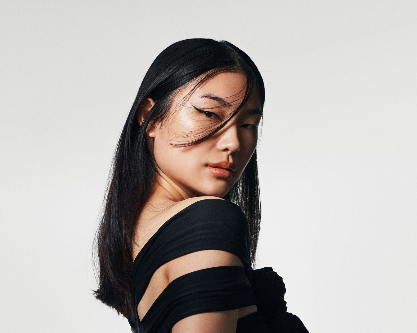 Primo piano di una modella asiatica con lunghi capelli neri lisci, trucco leggero e abito nero con spalle scoperte, che guarda indietro sopra la spalla.