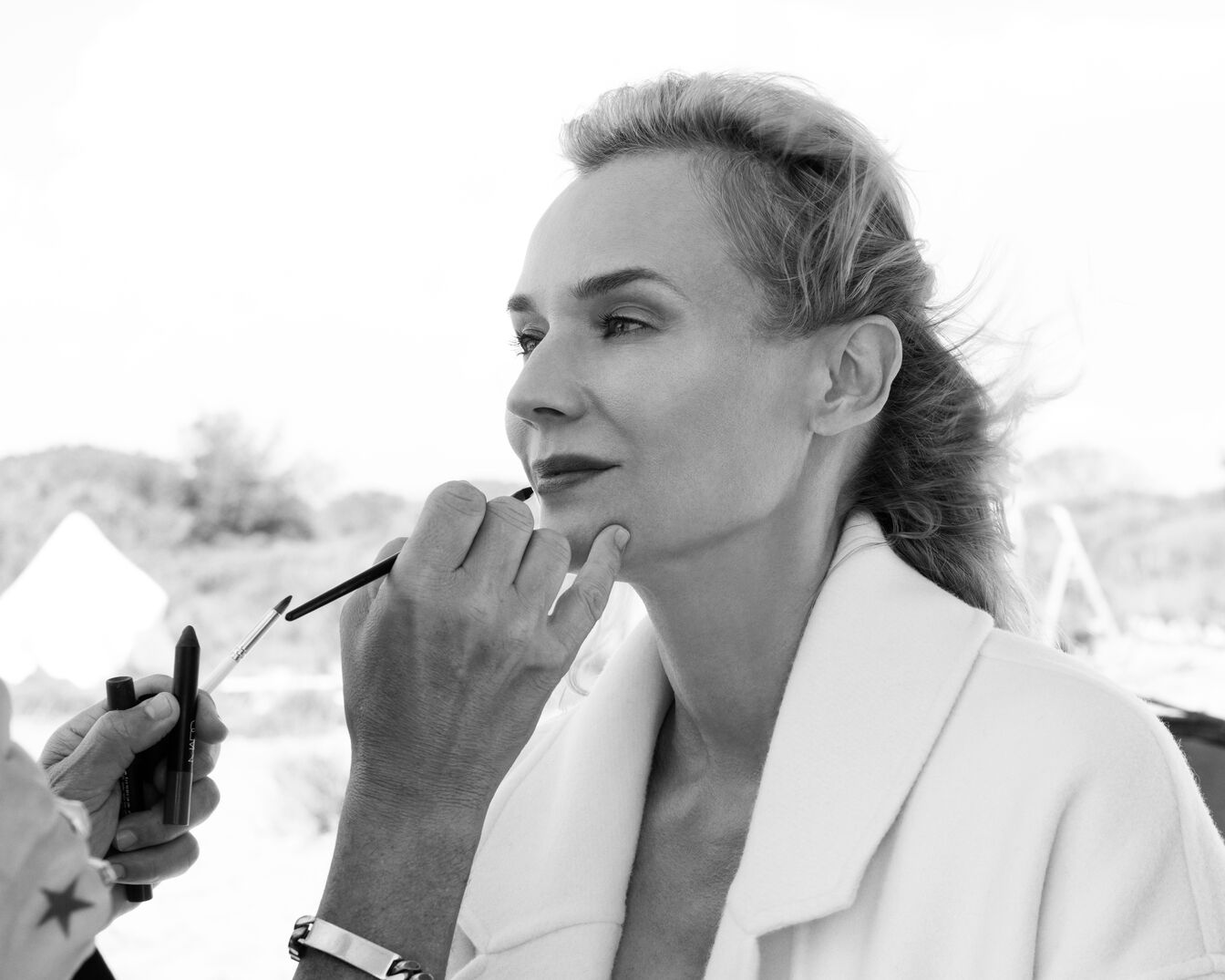 Czarno-białe zdjęcie kobiety, której wizażysta nakłada makijaż pędzlem, kobieta ma na sobie biały płaszcz.