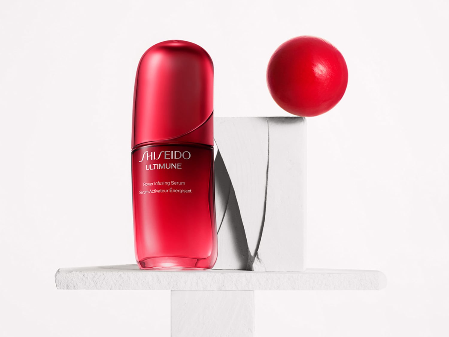 Flacone rosso di siero attivatore energetico Shiseido Ultimune su una piattaforma bianca, con una sfera rossa in equilibrio su un blocco.