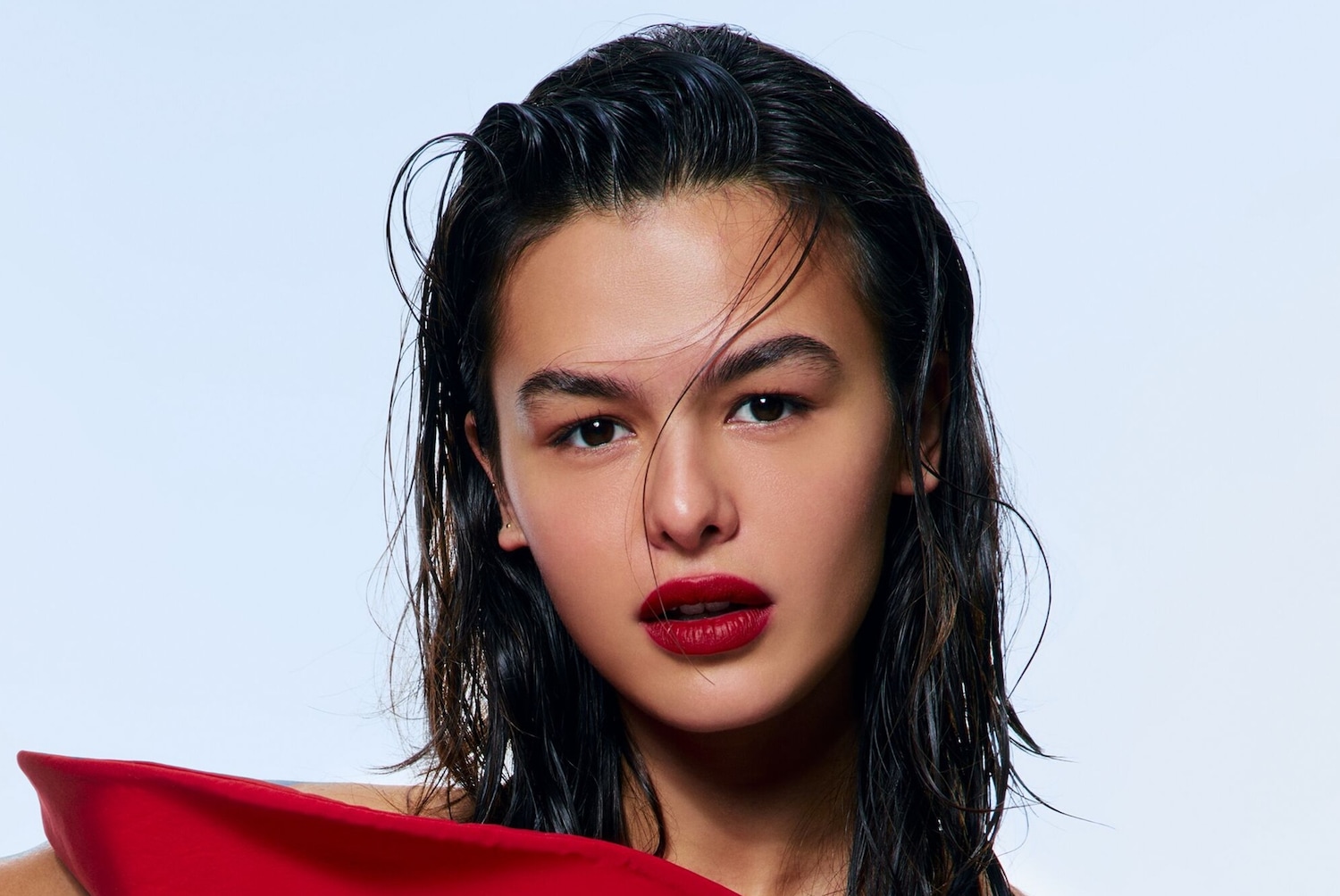 Portret kobiety z mokrymi, ciemnymi włosami i intensywnie czerwoną szminką, ubrana w czerwoną sukienkę na tle jasnego tła.
