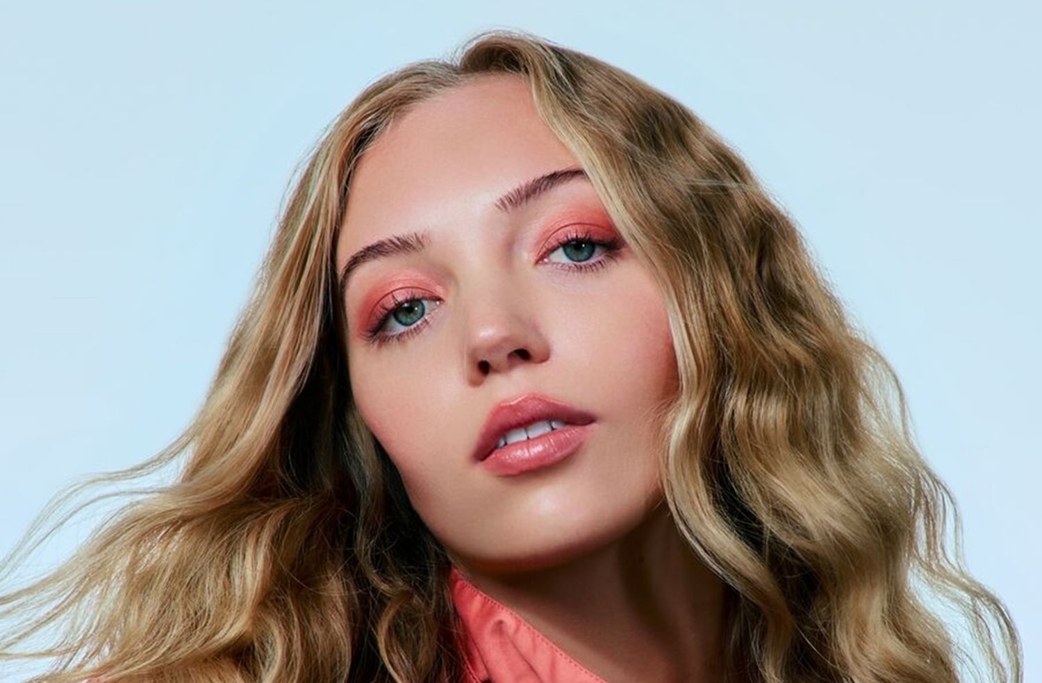 Portrét ženy s vlnitými blond vlasy a broskvovým očním make-upem, který ladí s jejím topem.