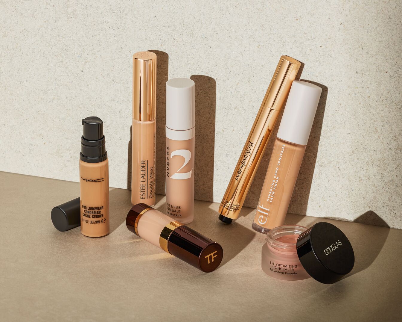 Una collezione di sei correttori di diverse marche, tra cui MAC, Estée Lauder, Morphe, Yves Saint Laurent, Elf e Douglas, sono disposti su una superficie chiara, con il correttore MAC Pro Longwear in una bottiglia di vetro trasparente con pompa nera, il correttore Estée Lauder Double Wear in un tubo beige con tappo dorato, il correttore Morphe Hide & Peek in un tubo beige con tappo bianco, il correttore Yves Saint Laurent in un tubo dorato, il correttore Elf Hydrating Camo in un tubo beige con tappo bianco e il correttore Douglas Eye Optimizing in un vasetto di vetro trasparente con tappo nero.