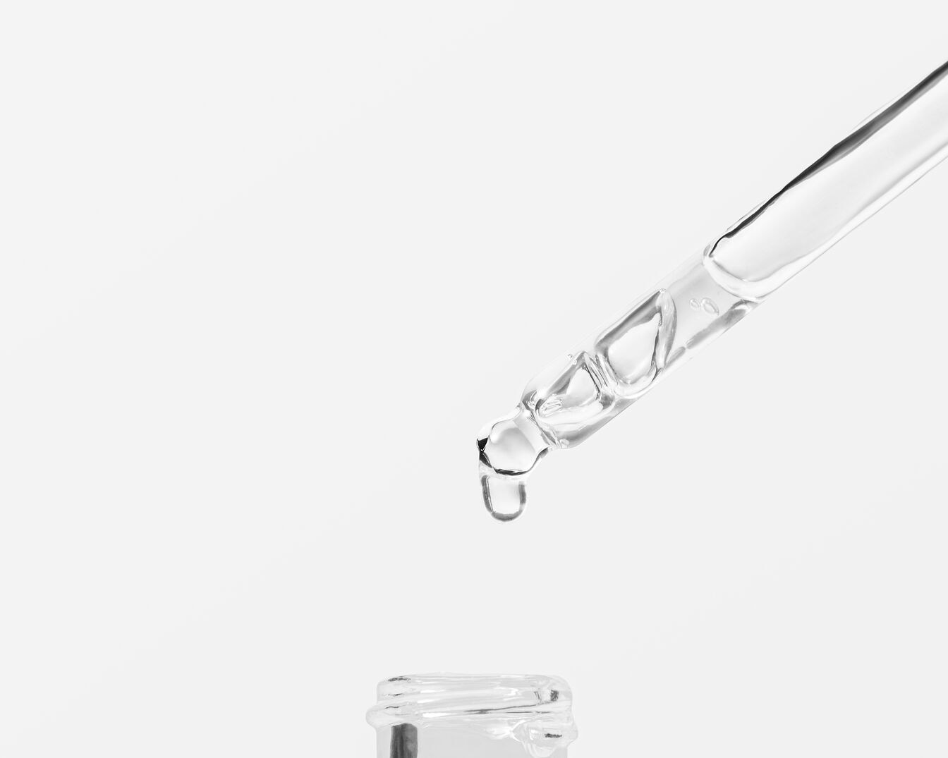Un compte-gouttes transparent laisse tomber une goutte de sérum clair dans un flacon ouvert, sur un fond blanc épuré.