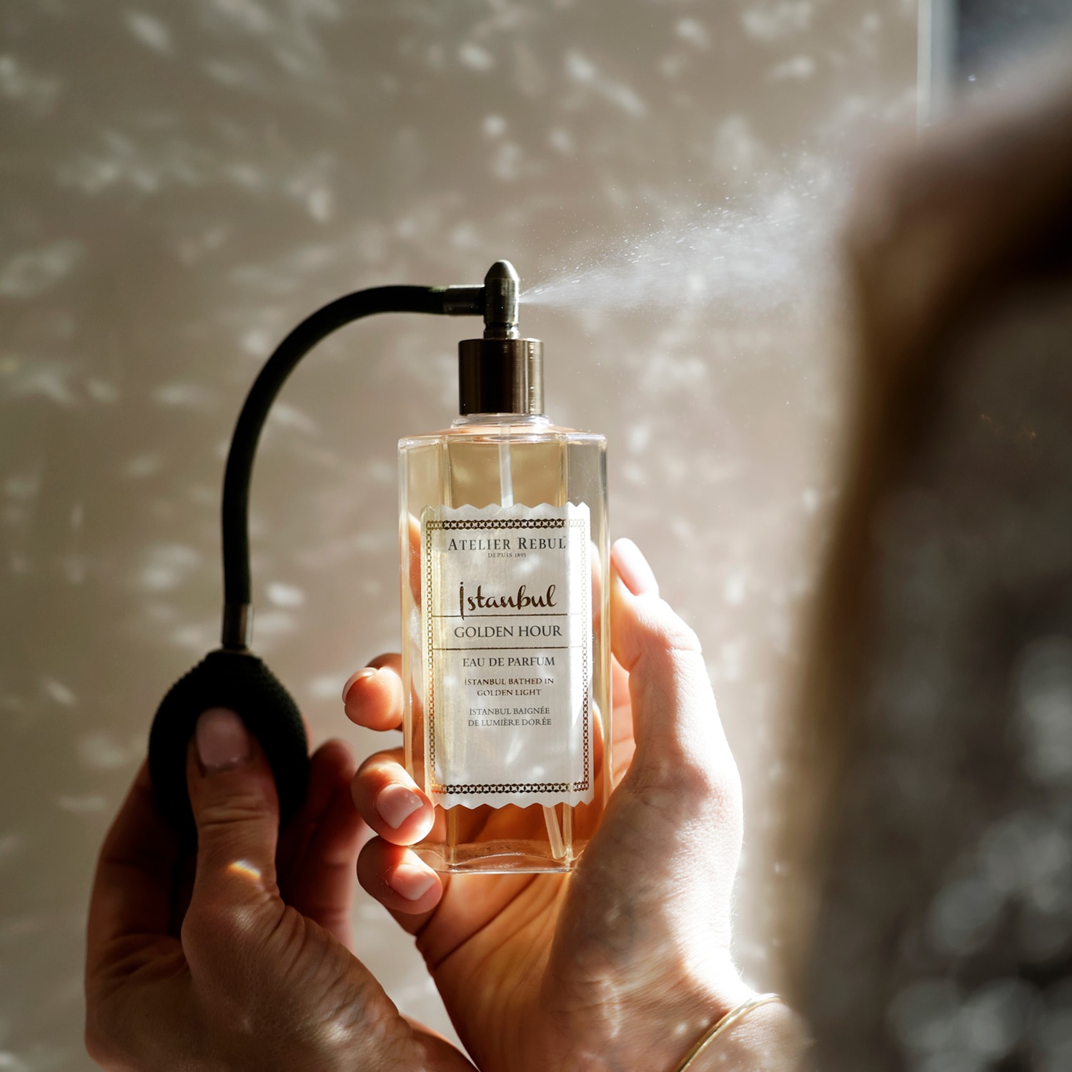 Een persoon spuit Atelier Rebul Istanbul Golden Hour Eau de Parfum, een heldere vloeistof in een vierkante glazen fles met een zwarte verstuiver, waarbij de fijne nevel zichtbaar is.