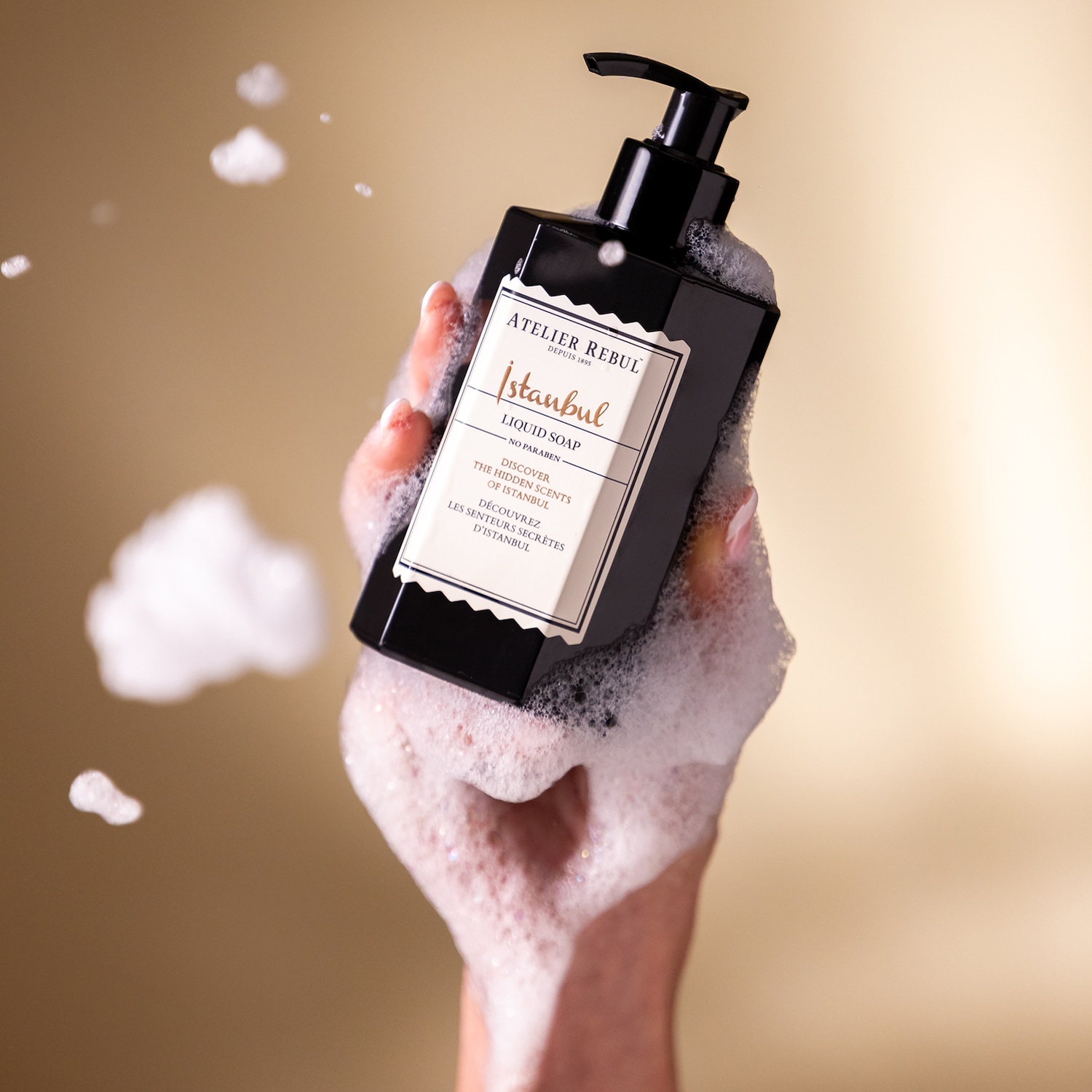 Een hand houdt een zwarte fles Atelier Rebul Istanbul vloeibare zeep vast, omringd door wit schuim, met de tekst "Discover the hidden scents of Istanbul" op het etiket.