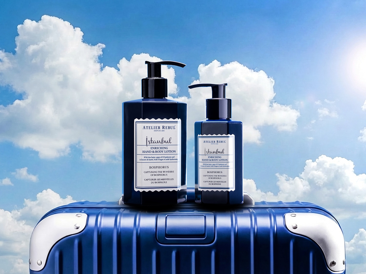 Twee donkerblauwe flessen Atelier Rebul Istanbul "Enriching Hand & Body Lotion" met zwarte pompen staan op een blauwe koffer tegen een helderblauwe lucht met witte wolken.