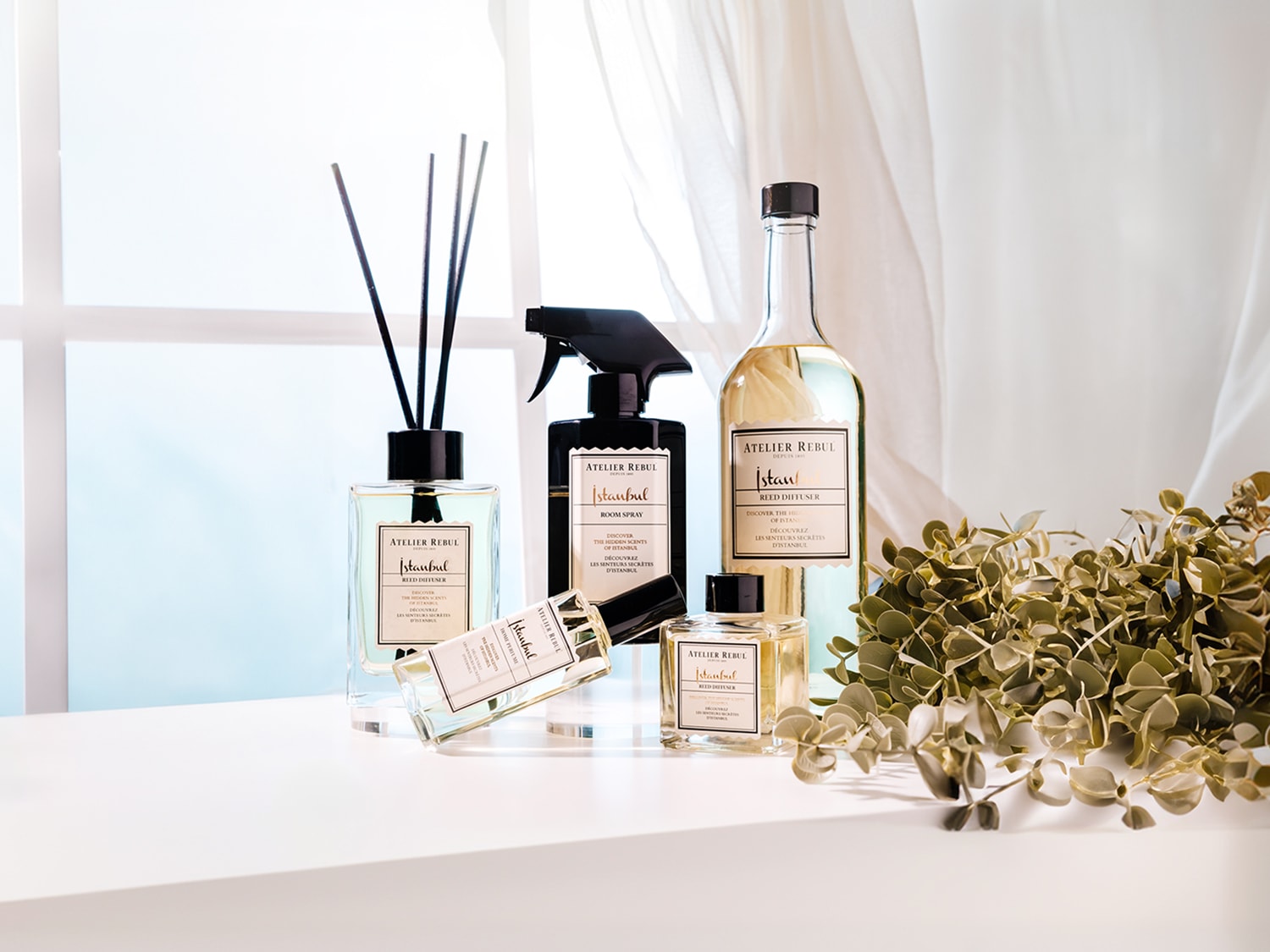 Een collectie Atelier Rebul Istanbul huisgeuren, waaronder een rietverspreider, kamerspray en parfum, staat op een witte ondergrond met een raam en gordijnen op de achtergrond, en een bosje groene bladeren aan de rechterkant.