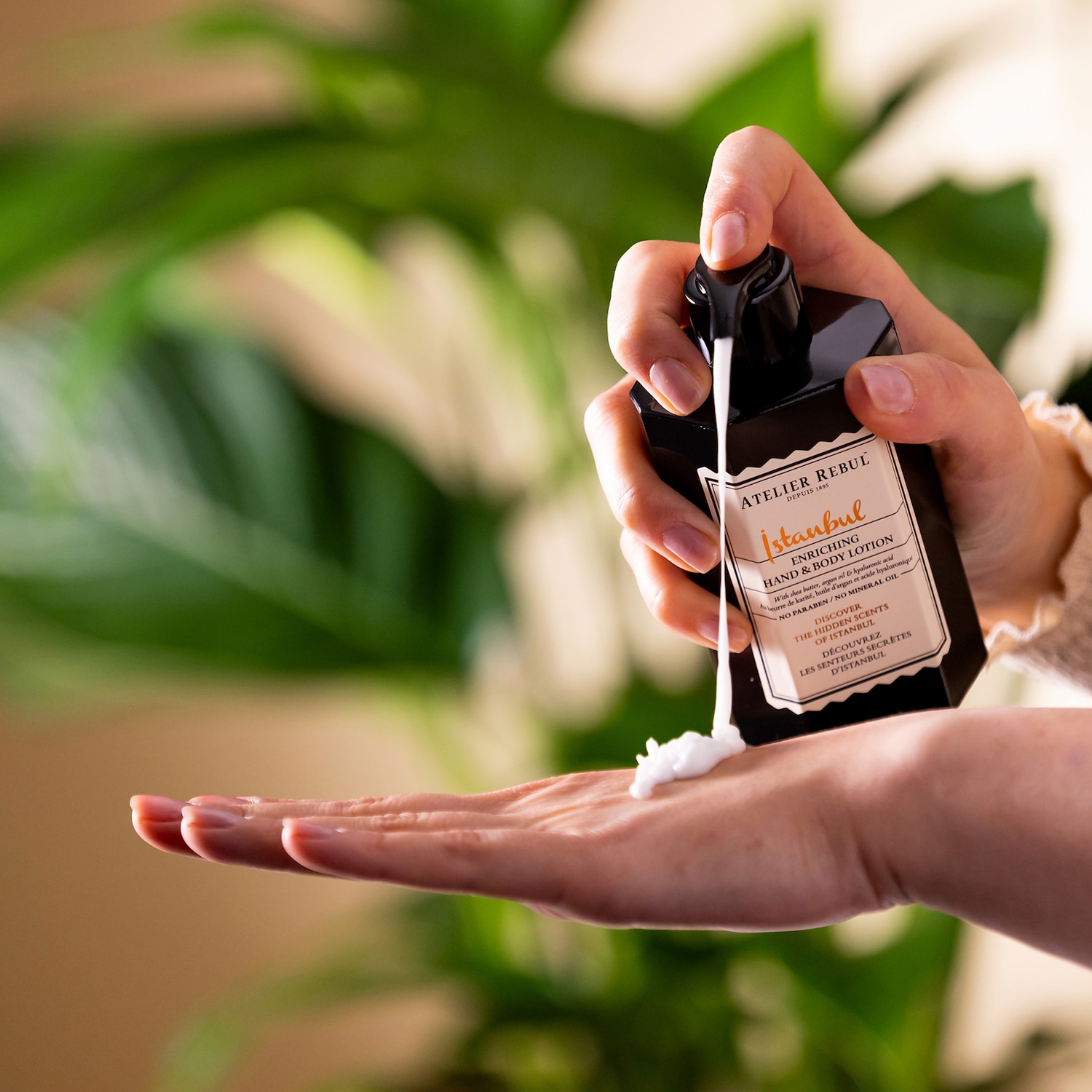 Een hand knijpt een zwarte fles Atelier Rebul Istanbul hand- en bodylotion, waardoor een witte crème in de open palm van de andere hand stroomt, met de tekst "ENRICHING HAND & BODY LOTION" en "NO PARABEN / NO MINERAL OIL" zichtbaar op het etiket.
