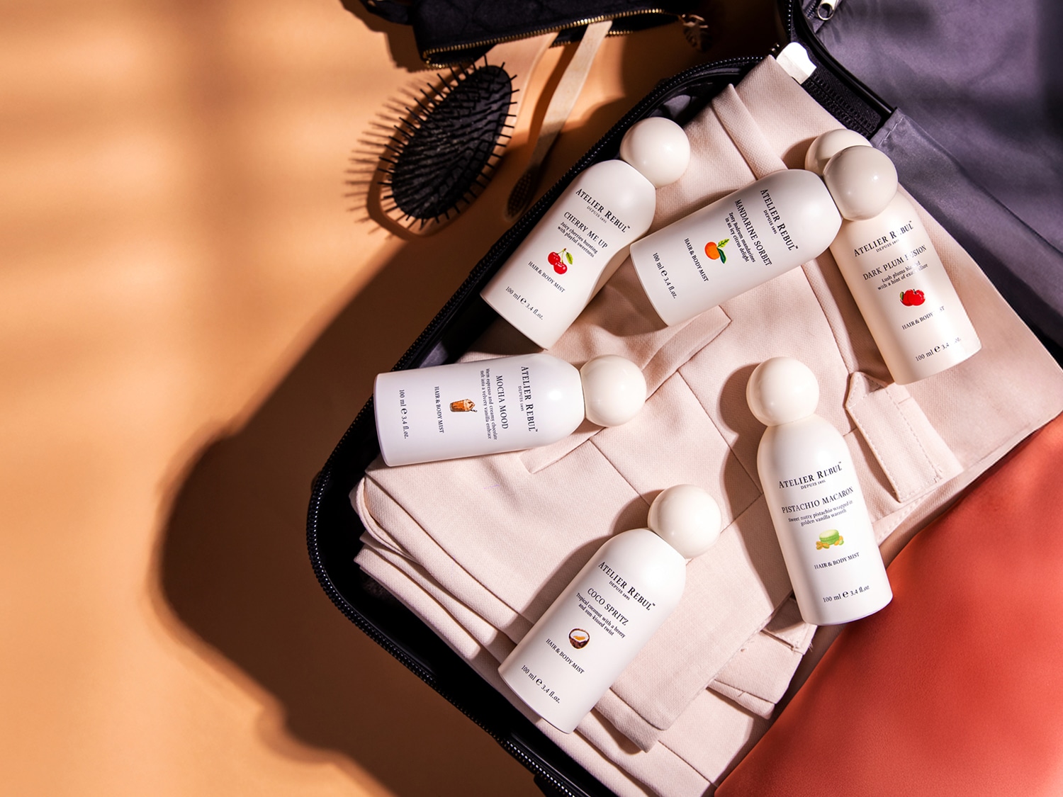 Zes witte flessen van Atelier Rebul Hair & Body Mist, elk met een inhoud van 100 ml, liggen in een open zwarte reistas met een haarborstel en kledingstukken.
