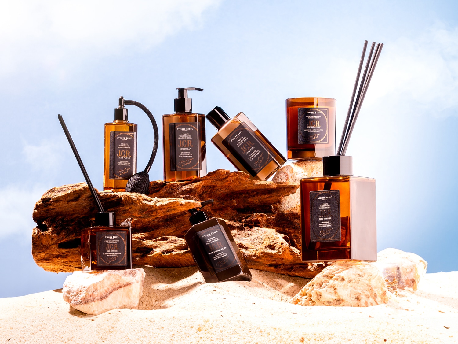 Een collectie Atelier Rebul J.C.R. producten, waaronder een parfumverstuiver, vloeibare zeep, douchegel, hand- en bodylotion, geurkaars en reed diffuser, gepresenteerd op zand en rotsen onder een blauwe lucht.
