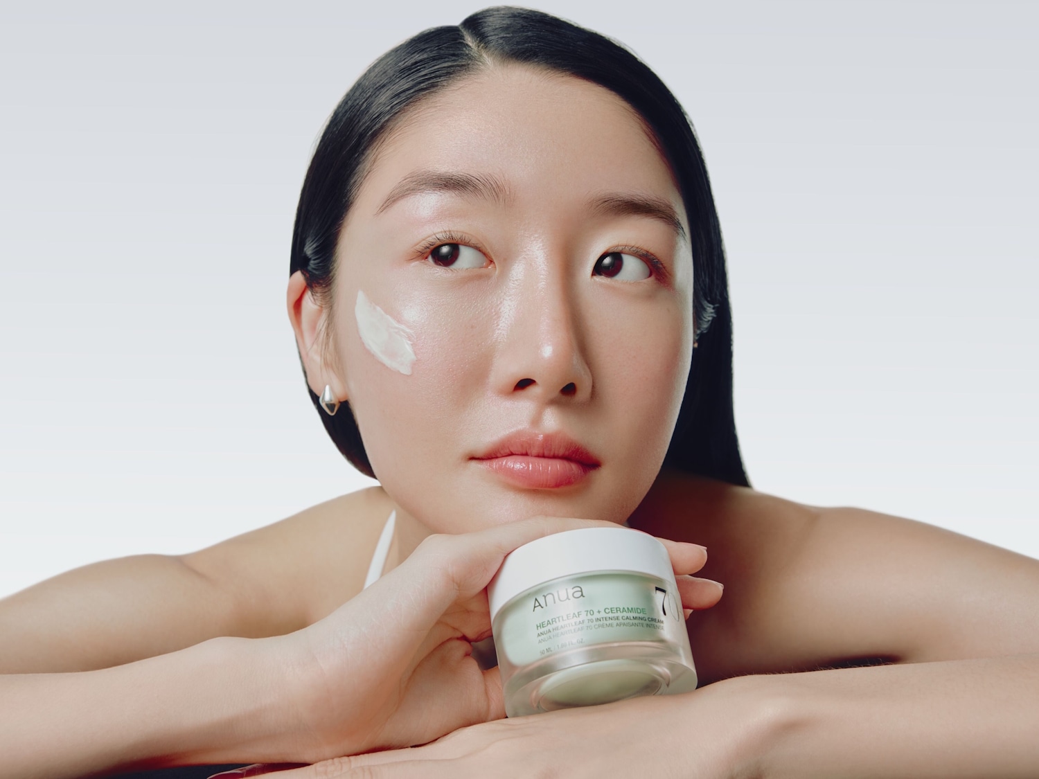 Eine Frau mit glänzender Haut hält einen Tiegel Anua Heartleaf 70 + Ceramide Intense Calming Cream in der Hand, während ein Klecks der Creme auf ihrer Wange zu sehen ist.