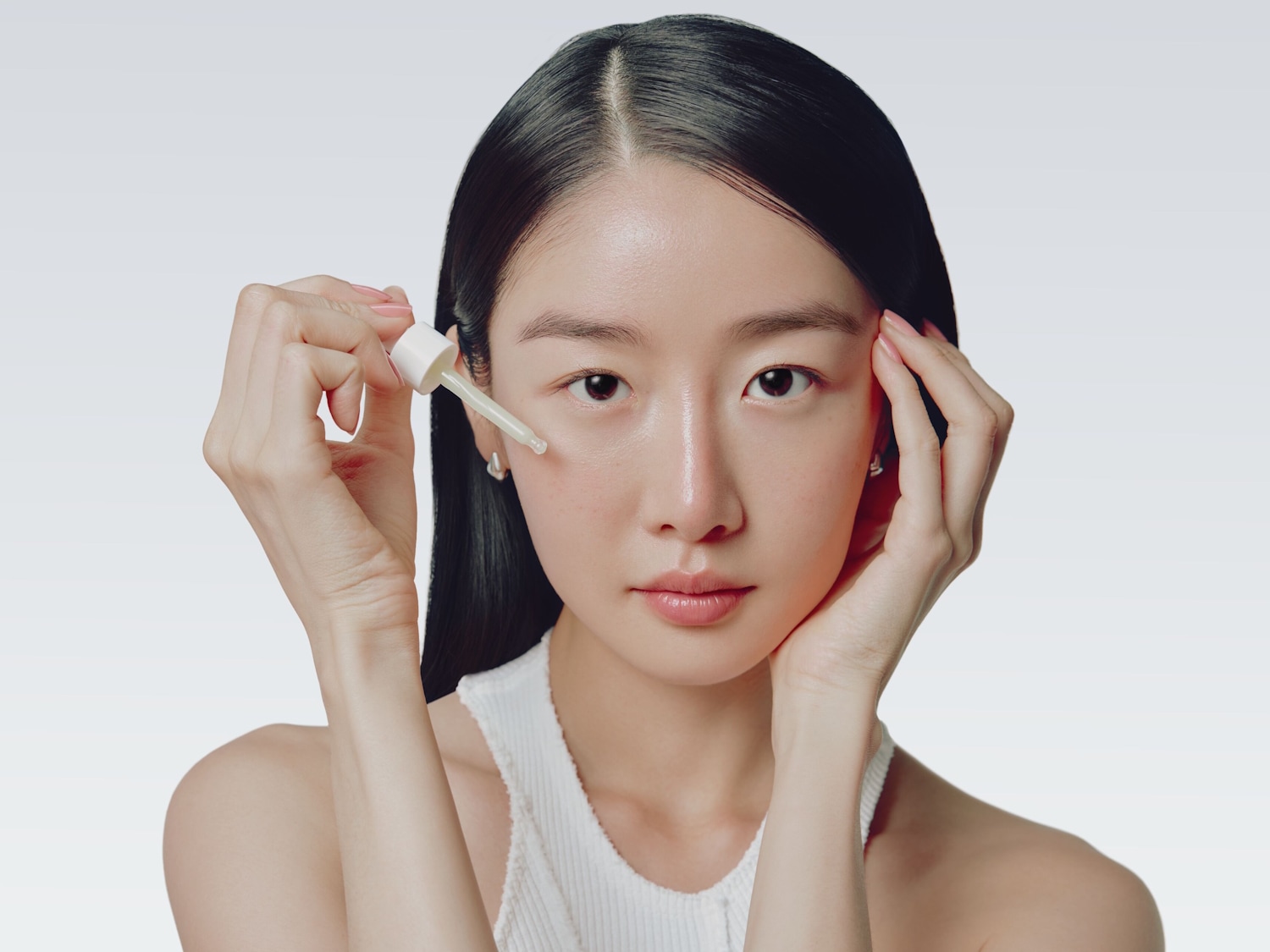 Eine Frau mit glänzender Haut trägt ein weißes Oberteil und hält eine Pipette mit einem Serum an ihr Gesicht, um die Anwendung eines Hautpflegeprodukts zu demonstrieren.
