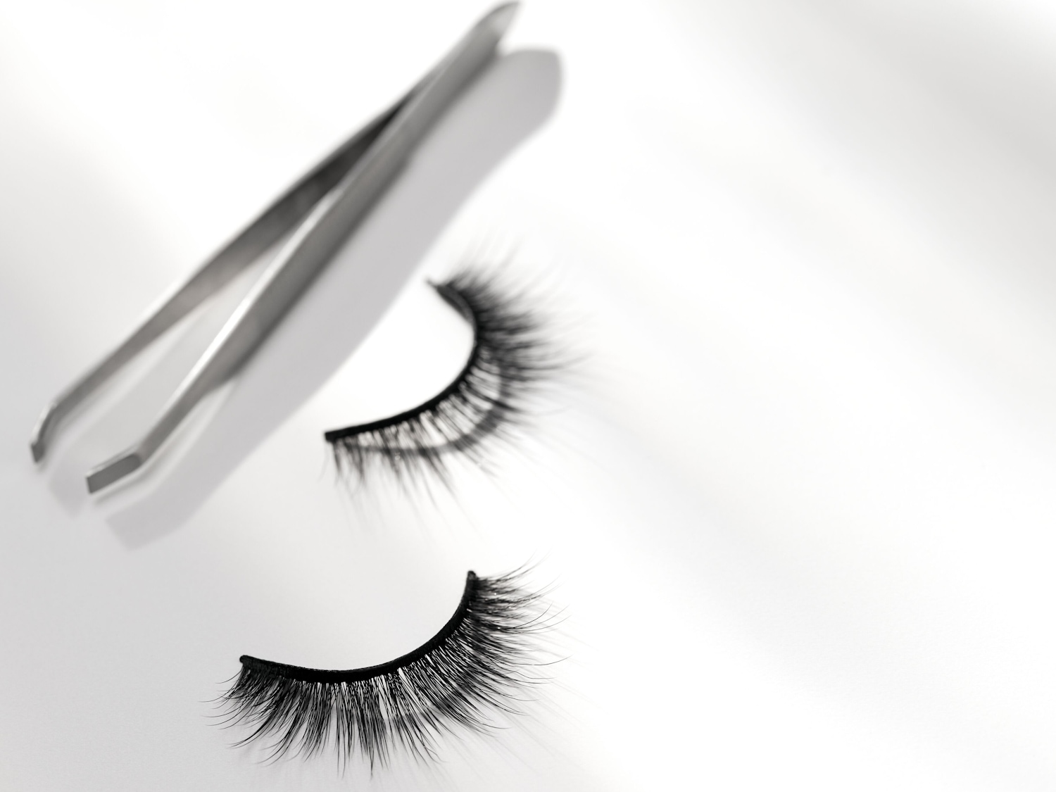 Paar zwarte valse wimpers en een zilveren pincet liggen op een witte achtergrond, klaar om je oogmake-up te perfectioneren.