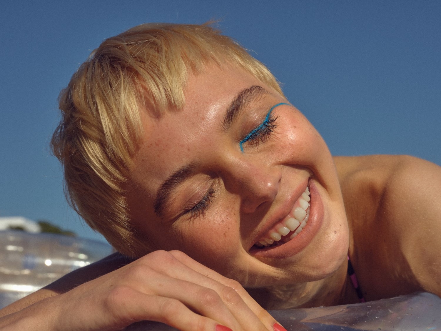 Een stralende vrouw met blond haar en blauwe eyeliner lacht terwijl ze ontspant tegen een helderblauwe achtergrond.