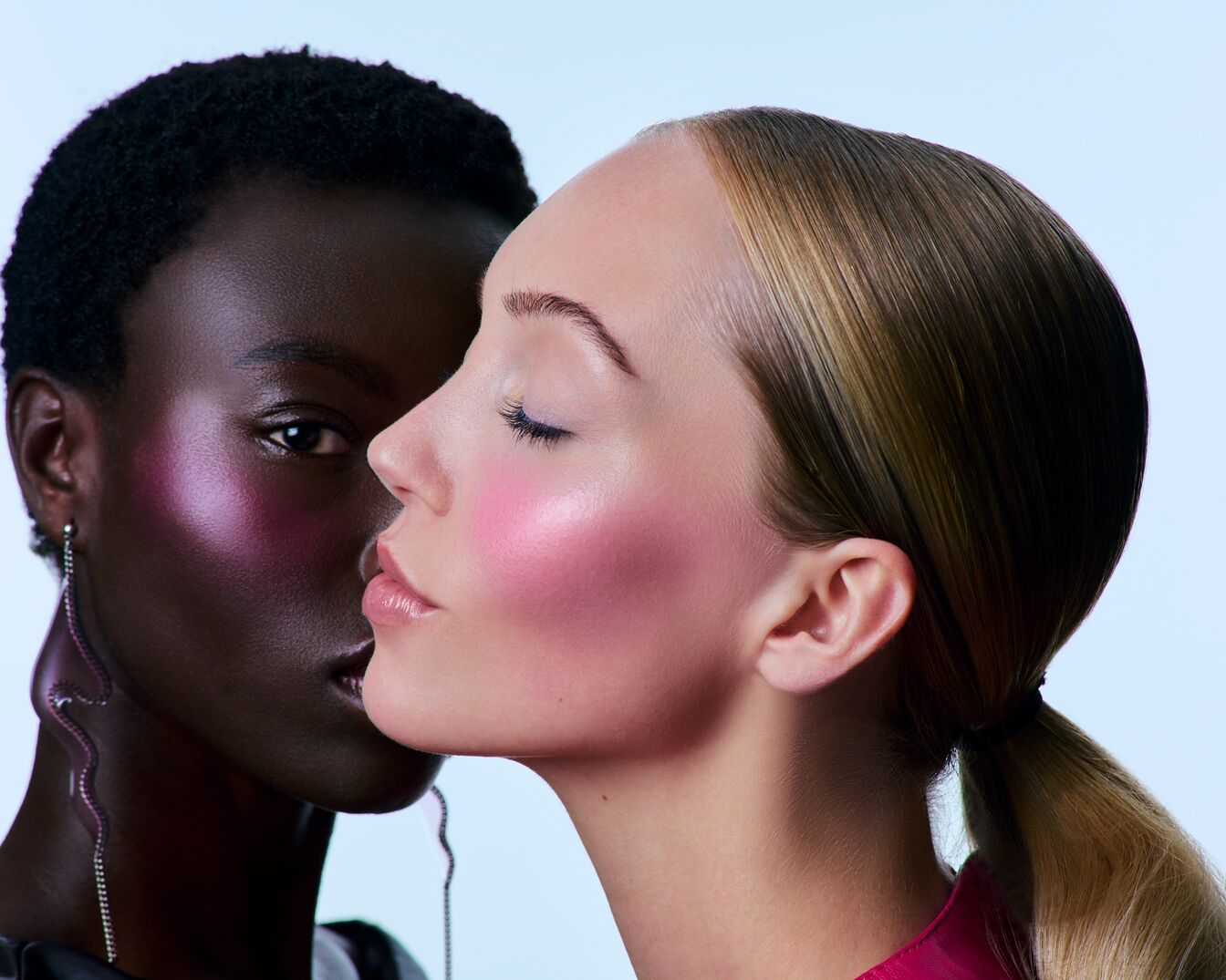 Nahaufnahme von zwei Frauen mit rosa Rouge, die eine mit dunkler Haut und Ohrring, die andere mit blonden Haaren im Pferdeschwanz.