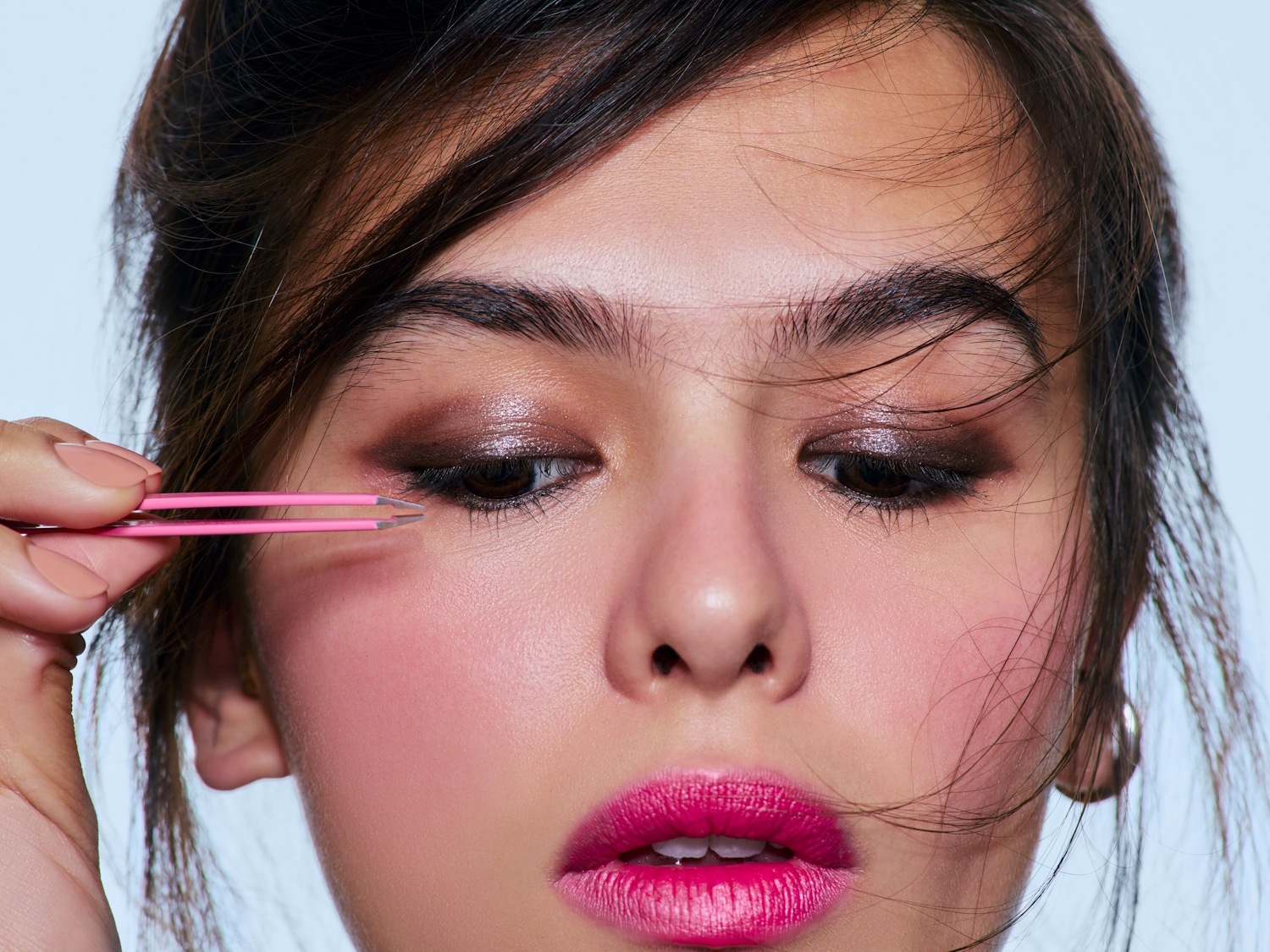 Nahaufnahme einer Frau, die eine pinkfarbene Pinzette an ihre Augen hält, mit schimmerndem Lidschatten und pinkfarbenem Lippenstift.