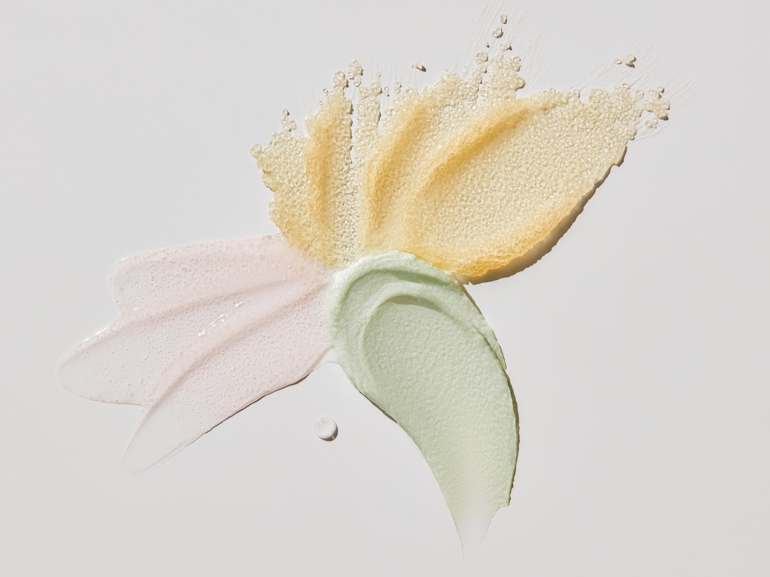 Verschillende texturen en kleuren van huidverzorgingsproducten, waaronder een roze lotion, een groene crème, en een gouden scrub, kunstzinnig gerangschikt op een witte achtergrond.