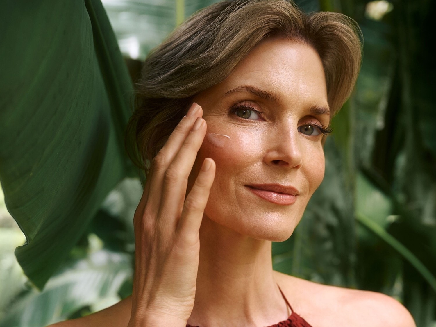 Eine Frau mit hellbraunem Haar und einem roten Oberteil trägt eine Feuchtigkeitscreme auf ihre Wange auf, während sie vor einem Hintergrund aus grünen Blättern posiert.