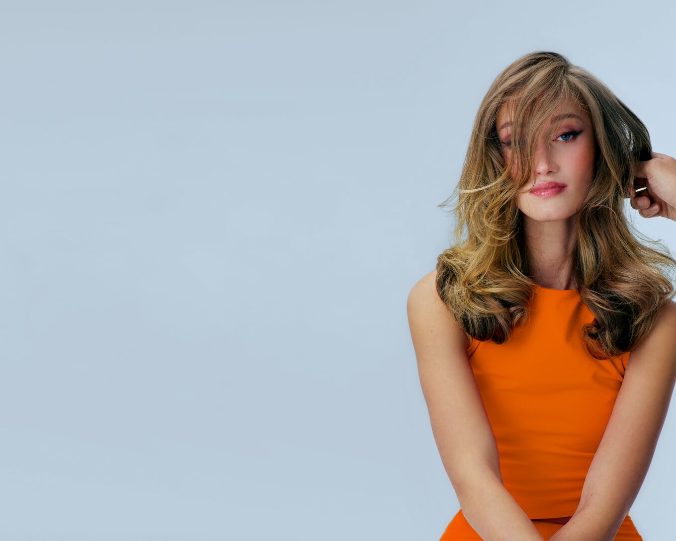 Une femme aux longs cheveux ondulés châtain clair, portant un haut orange vif, pose avec une main ajustant ses cheveux devant un fond bleu clair.