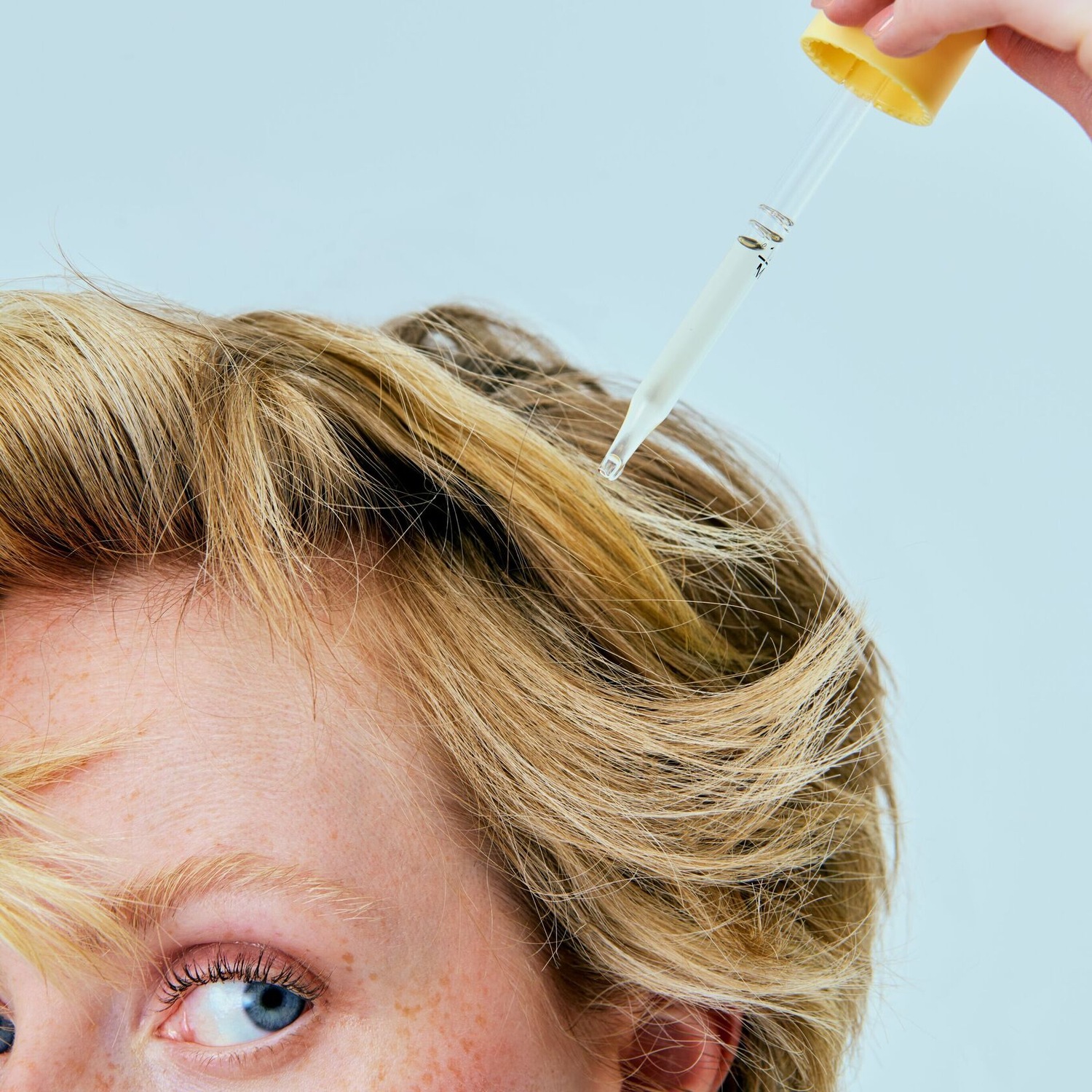 Gros plan sur une personne aux cheveux blonds recevant un traitement avec un compte-gouttes, mettant en évidence l'application précise d'un sérum capillaire pour des soins ciblés.