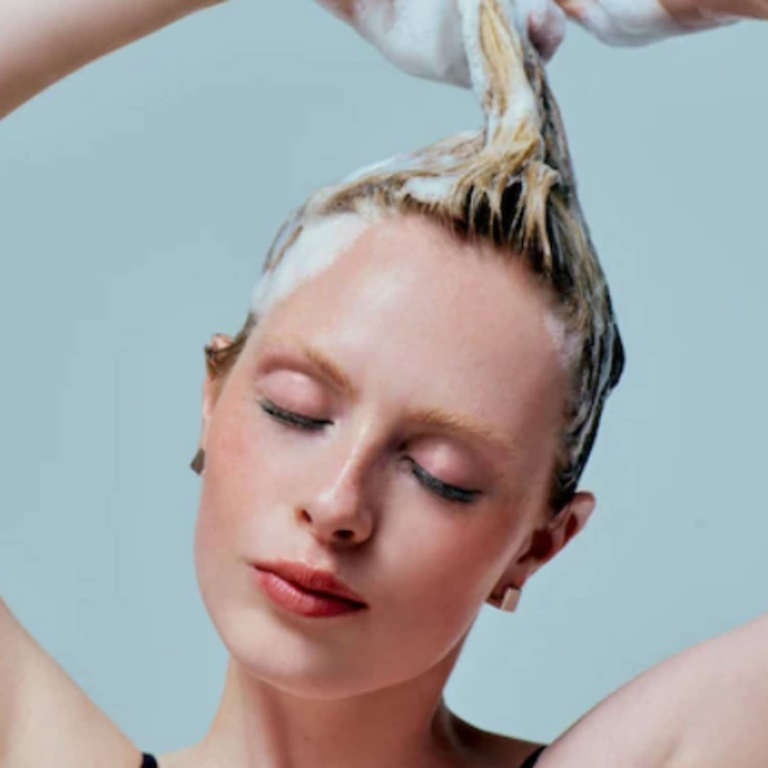 Une femme aux cheveux blonds savonne ses cheveux avec les yeux fermés, créant une mousse blanche et abondante.