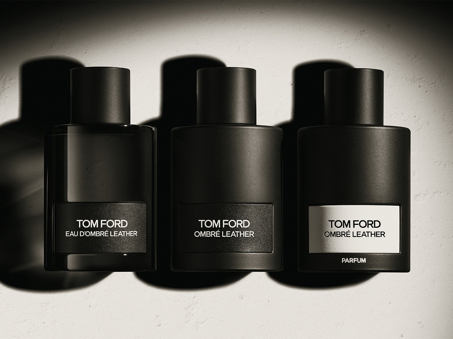Tres frascos de perfume Tom Ford Ombré Leather en diferentes concentraciones: Eau d'Ombré Leather, Ombré Leather y Ombré Leather Parfum, cada uno con un diseño elegante y minimalista en tonos oscuros.