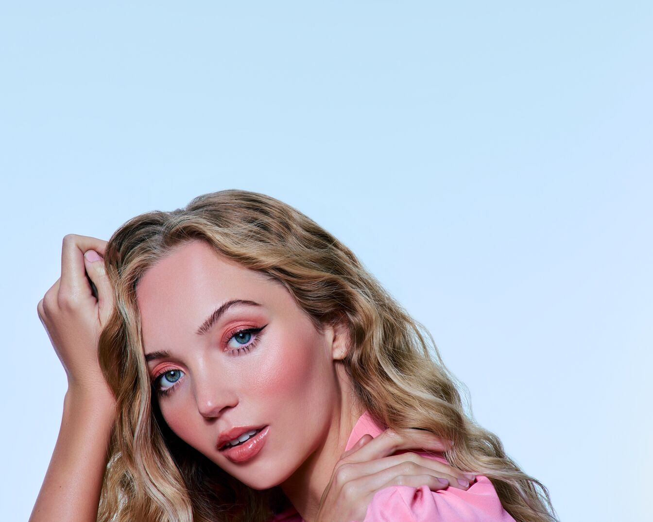 Portrait d'une femme aux cheveux blonds ondulés, portant un chemisier rose et un maquillage rose assorti sur les yeux, les joues et les lèvres, le tout sur un fond bleu clair.