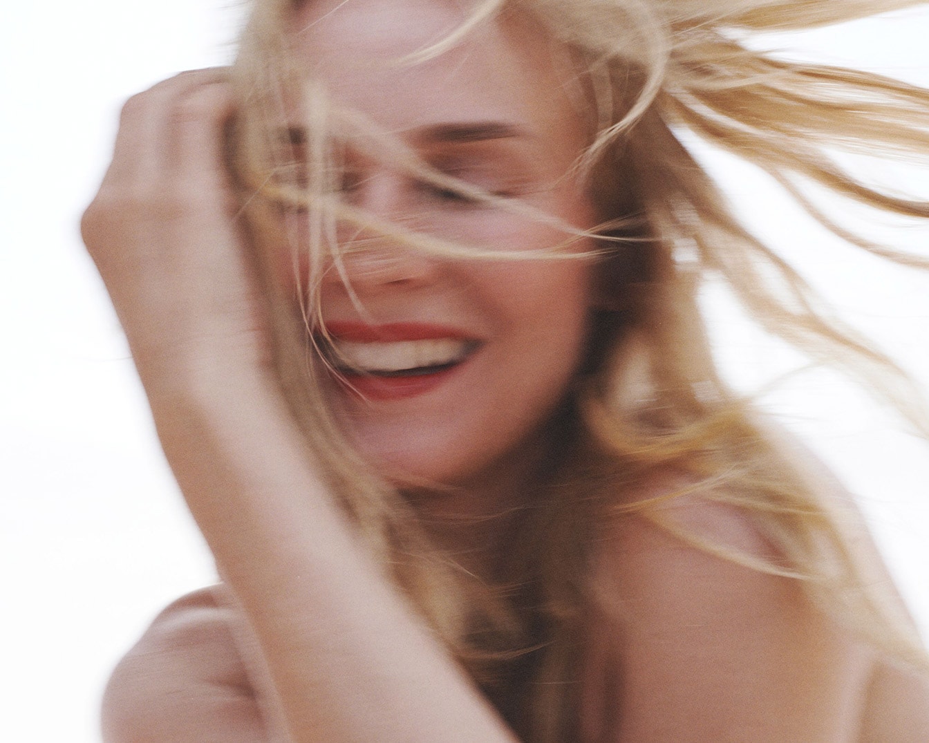 Une femme blonde, les cheveux au vent, sourit avec un rouge à lèvres rouge éclatant.