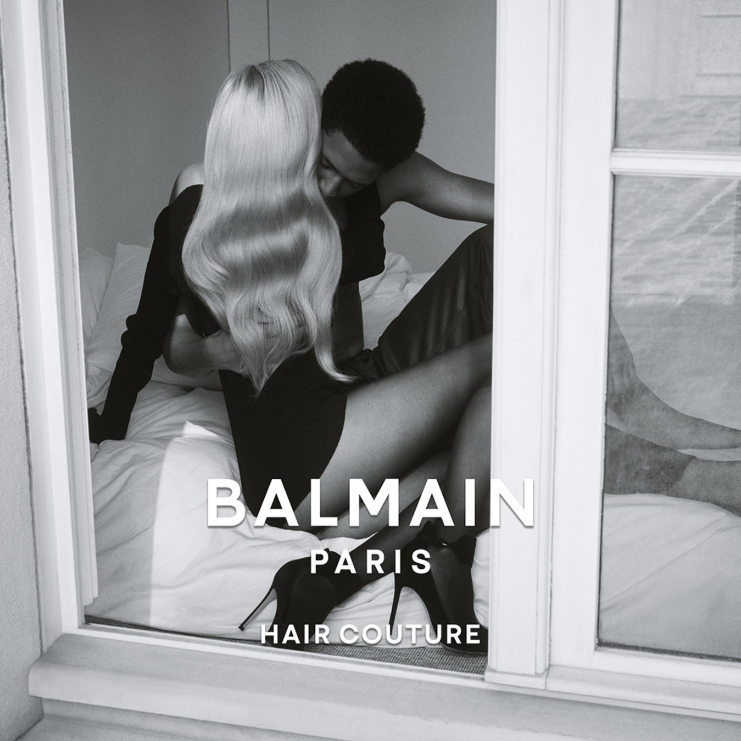 Černobílý snímek páru v objetí na posteli, viděný oknem, s nápisem BALMAIN PARIS HAIR COUTURE.