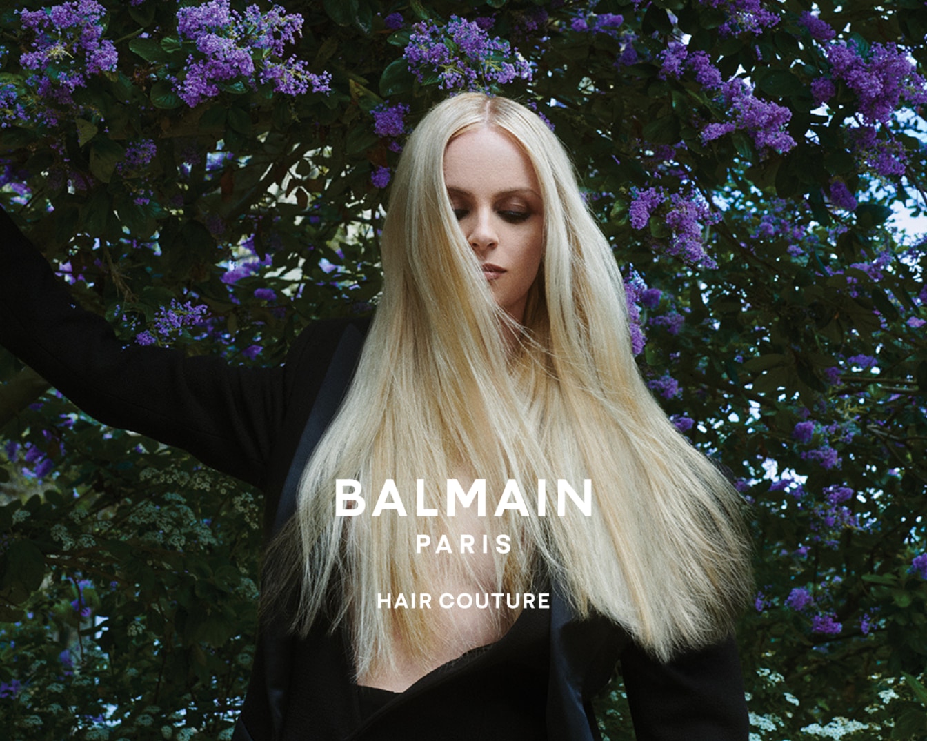 Na obrázku je blond modelka s dlhými vlasmi, oblečená v čiernom saku, stojí pred kvitnúcim kríkom a prezentuje Balmain Paris Hair Couture.