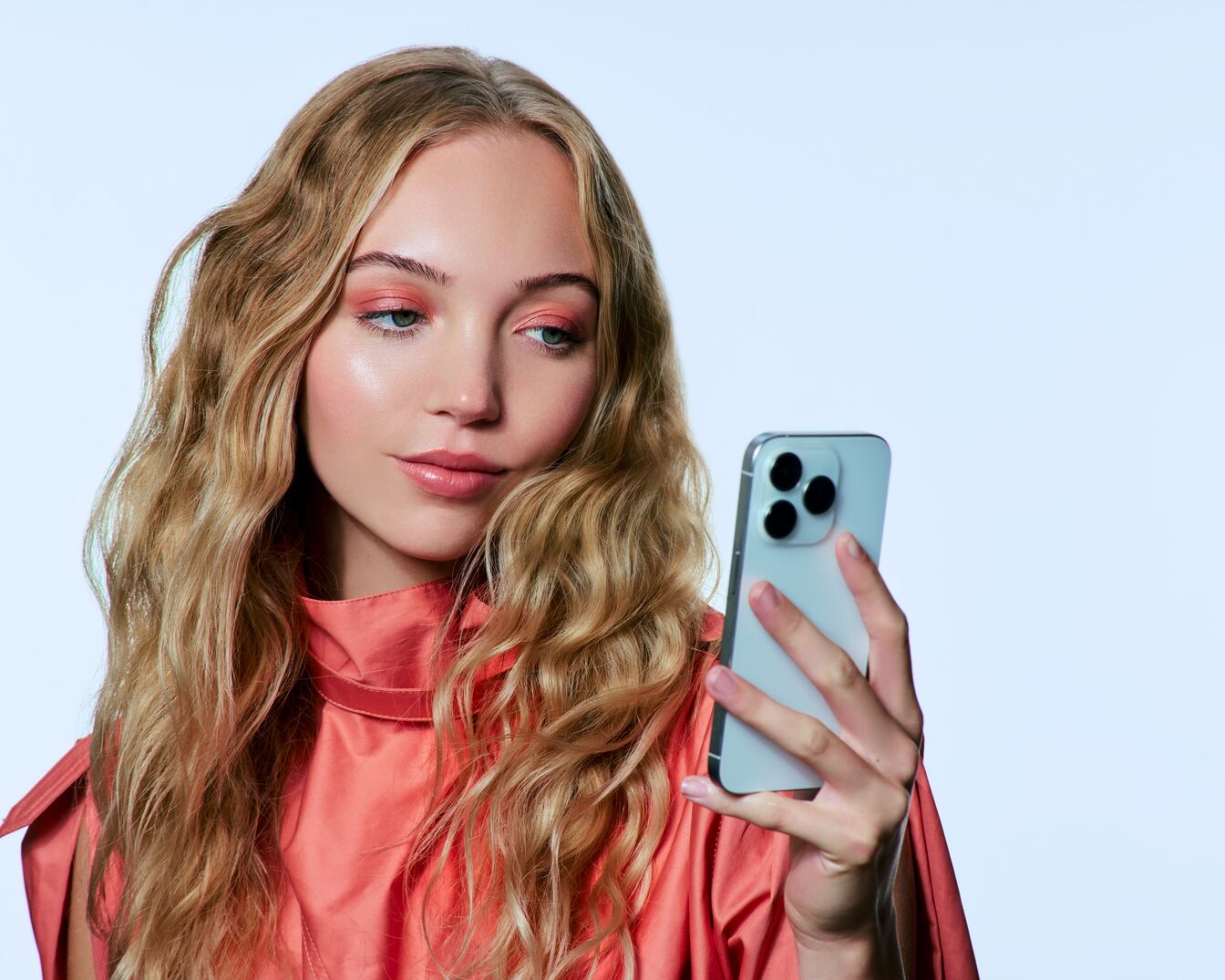 Blonde Frau mit Pfirsich-farbenem Lidschatten und passendem Oberteil, die ein hellblaues Smartphone in der Hand hält.