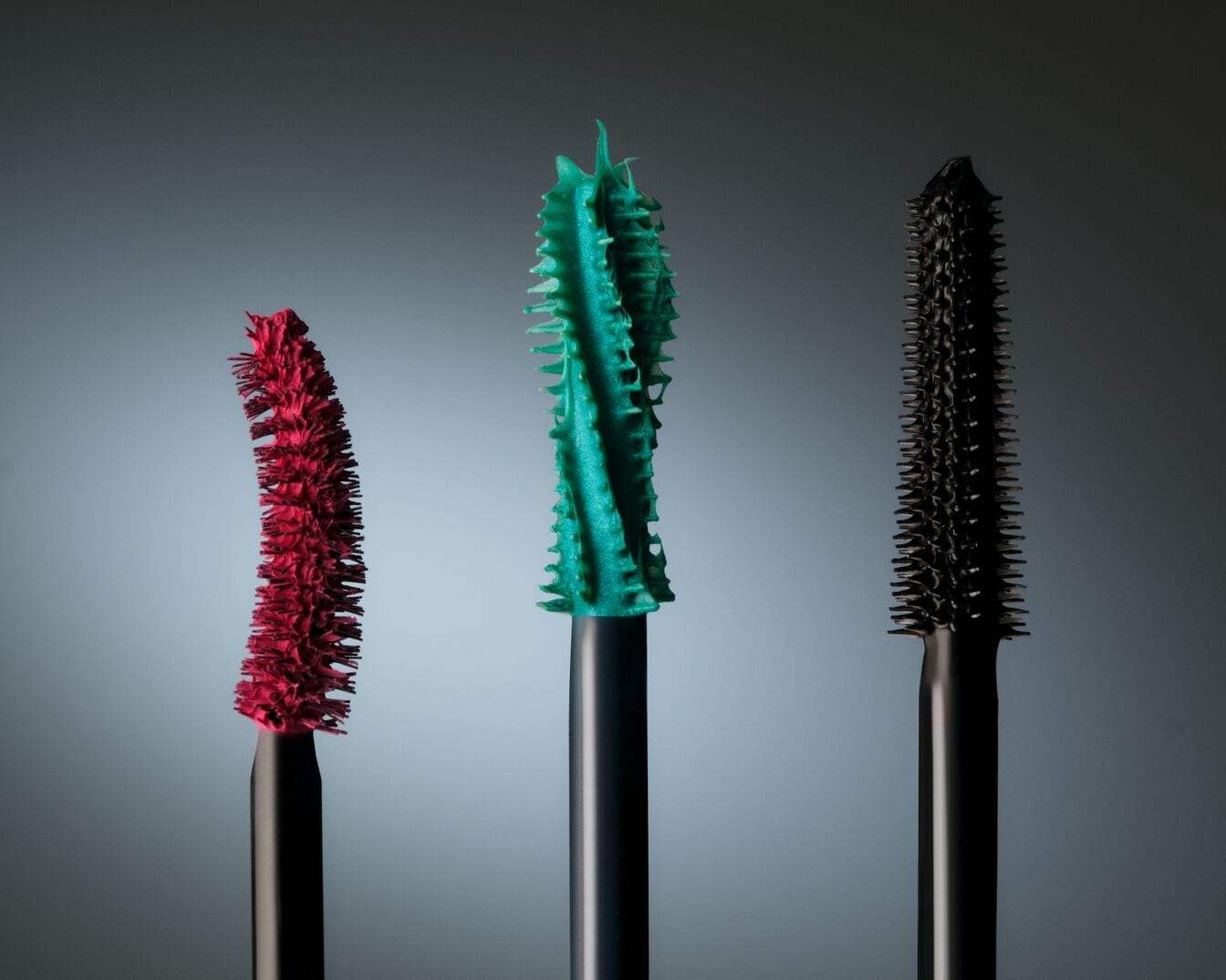 Gros plan sur trois brosses de mascara colorées : rouge, turquoise et noire, chacune avec une texture unique pour différents effets.