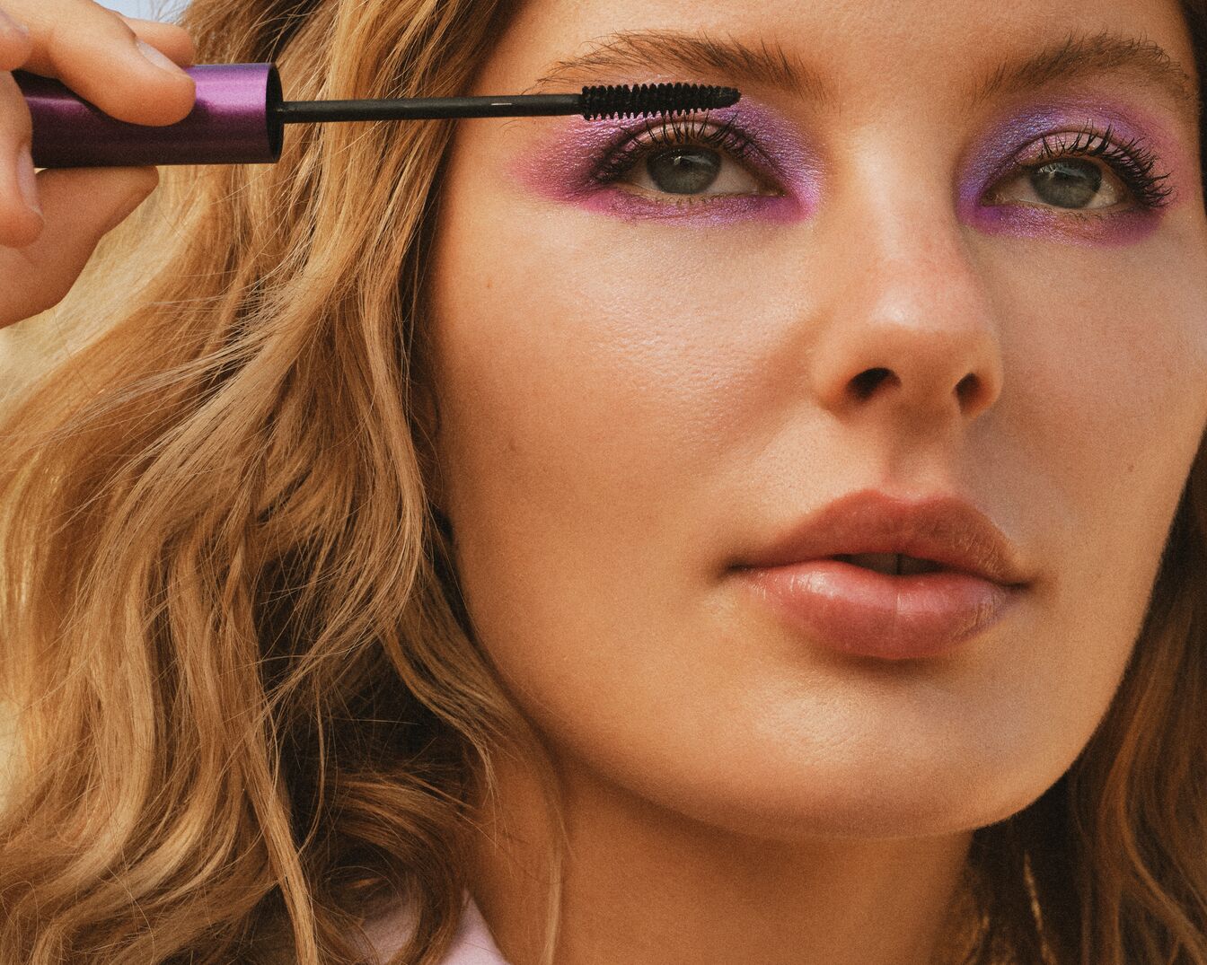 Gros plan sur une femme appliquant du mascara violet sur ses cils, avec un fard à paupières violet scintillant et des lèvres brillantes.
