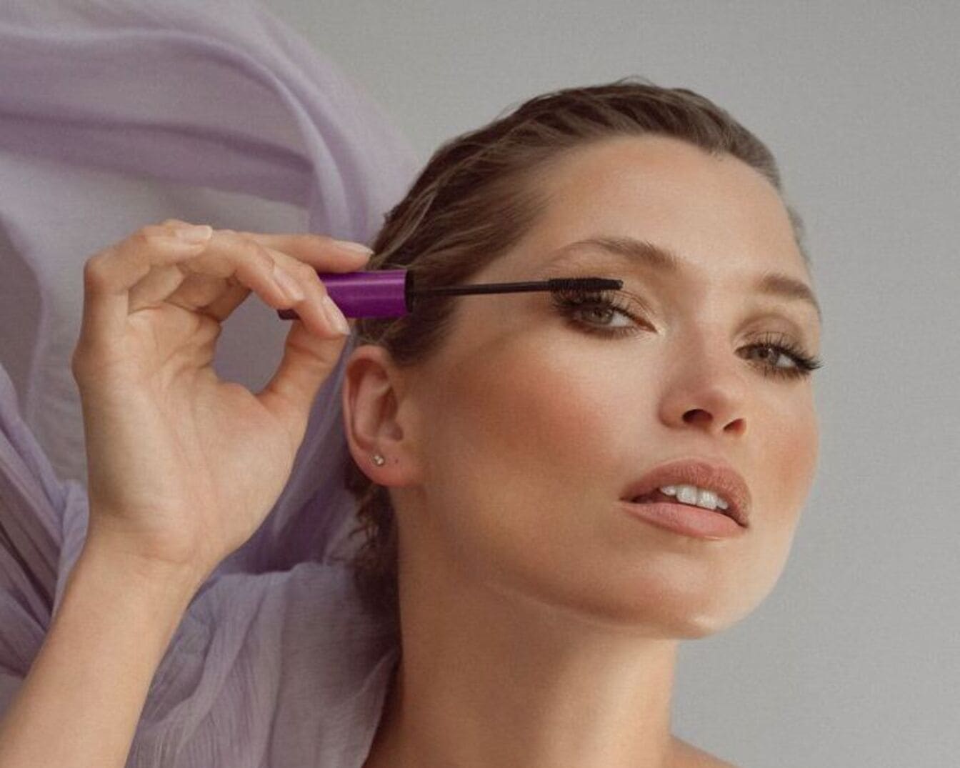 Une femme applique du mascara noir à l'aide d'une brosse sur ses cils, le mascara est dans un tube violet.