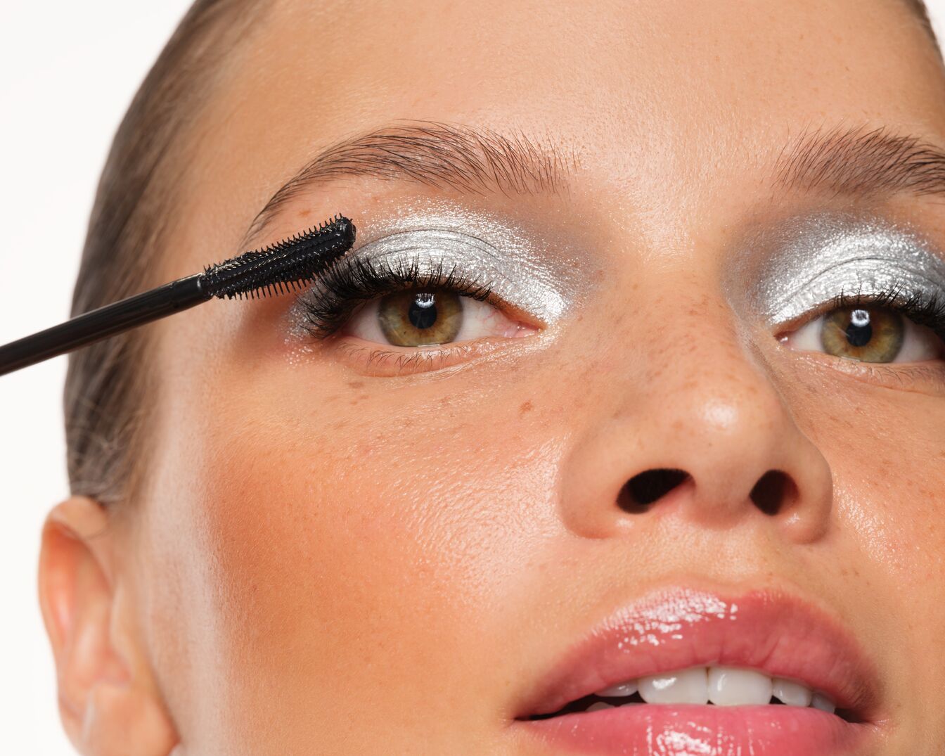 Gros plan d'une femme appliquant du mascara noir sur ses cils, avec un fard à paupières argenté scintillant qui illumine son regard.
