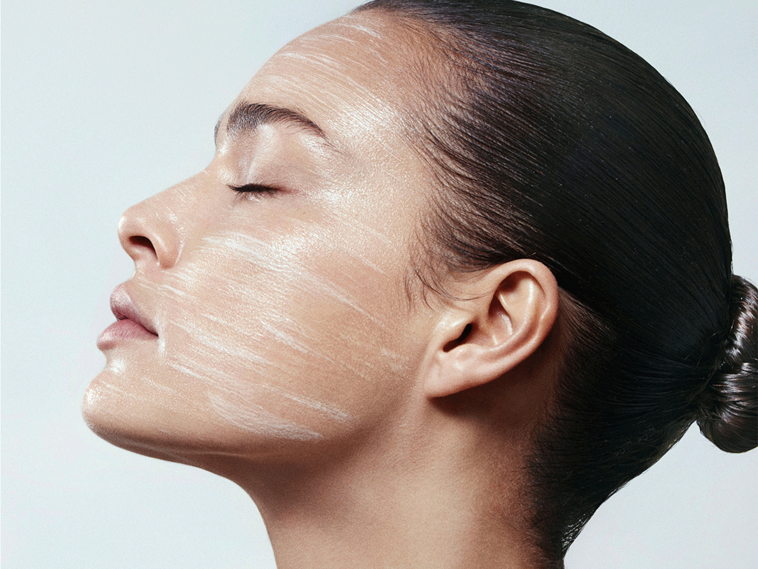 Profil d'une femme avec les yeux fermés et un masque blanc crémeux appliqué sur son visage, ses cheveux noirs tirés en chignon.