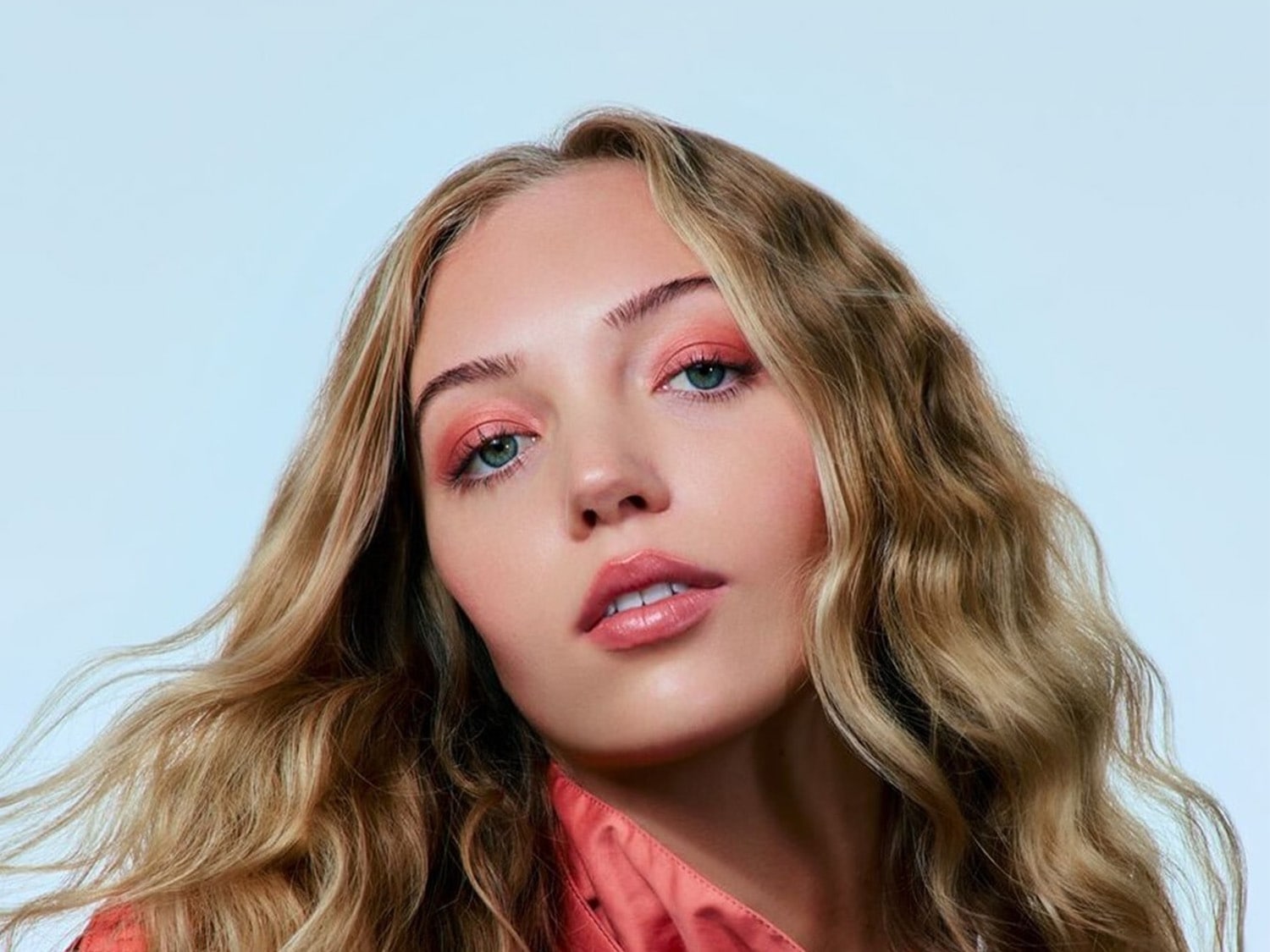 Een close-up van een vrouw met blond haar, perzikroze oogschaduw en glanzende lippenstift, die een zachte en frisse make-uplook laat zien.