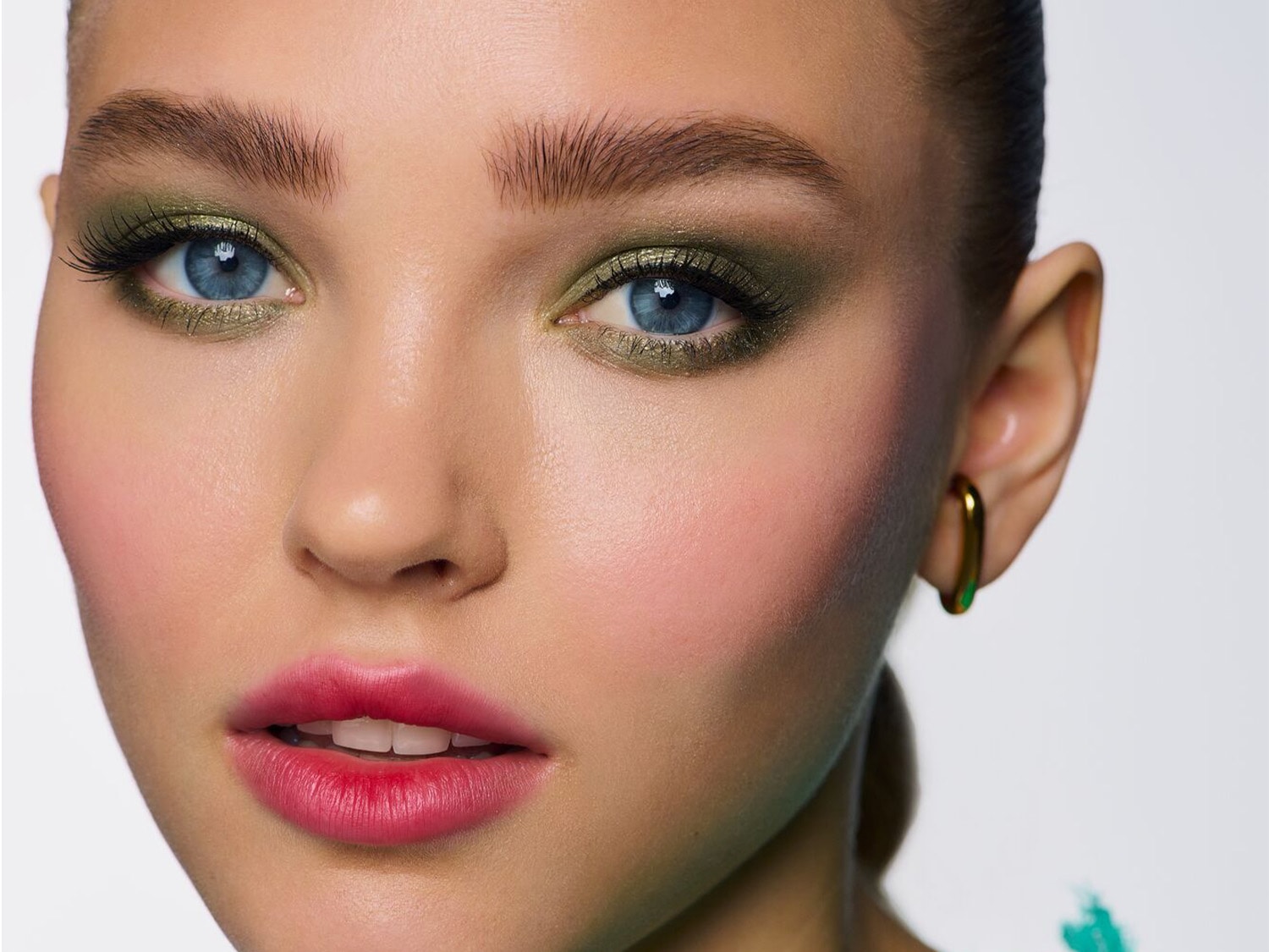 Close-up van een vrouw met blauwe ogen, groene oogschaduw en roze lippenstift, die een gouden oorbel draagt.