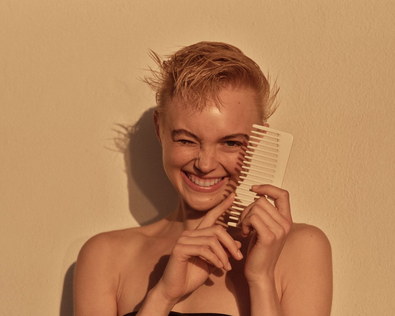 Een lachende vrouw met kort blond haar houdt een witte kam voor haar gezicht, terwijl ze knipoogt en haar tanden laat zien.