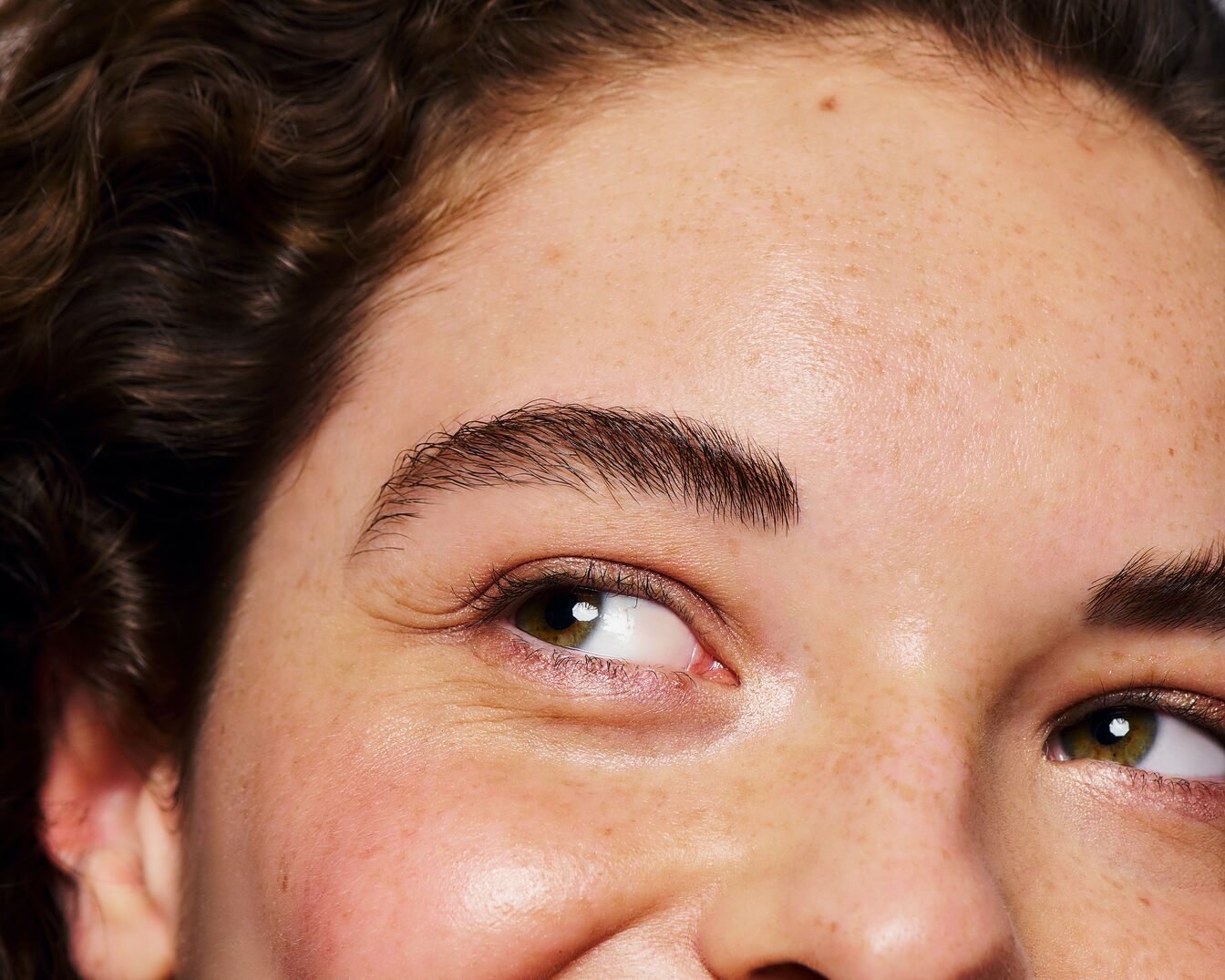 Close-up van een vrouw met bruin krullend haar, groene ogen en volle wenkbrauwen, lachend met sproeten op haar gezicht.