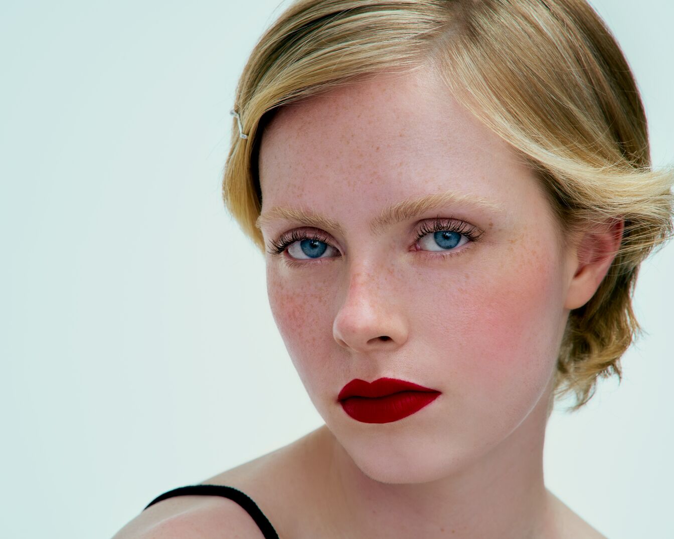 Een close-up van een vrouw met blauwe ogen, sproeten en felrode lippenstift, haar blonde haar is kort en golvend.