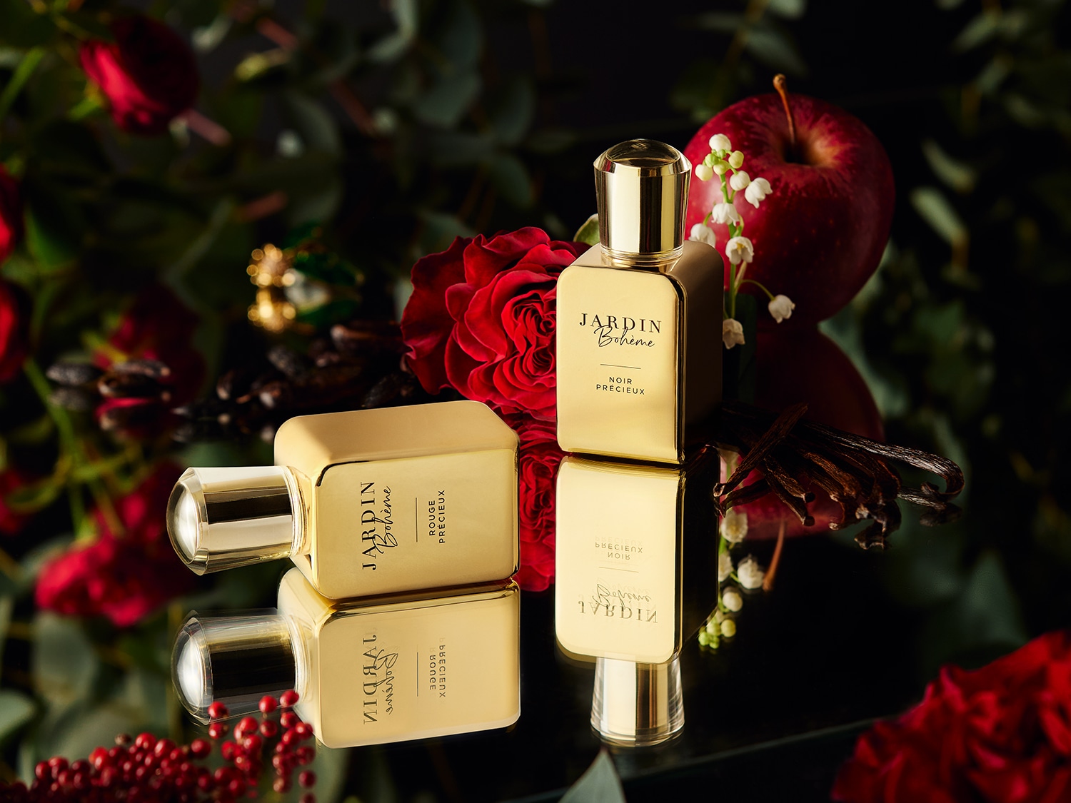 Zwei goldene Parfümflaschen von Jardin Bohème, eine mit der Aufschrift „Rouge Précieux“ und die andere mit „Noir Précieux“, umgeben von roten Rosen, einem Apfel und Vanilleschoten.
