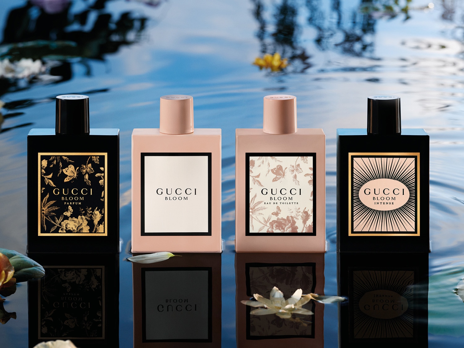 Čtyři flakony parfémů Gucci Bloom v různých variantách, umístěné na vodní hladině s odrazy a lekníny.