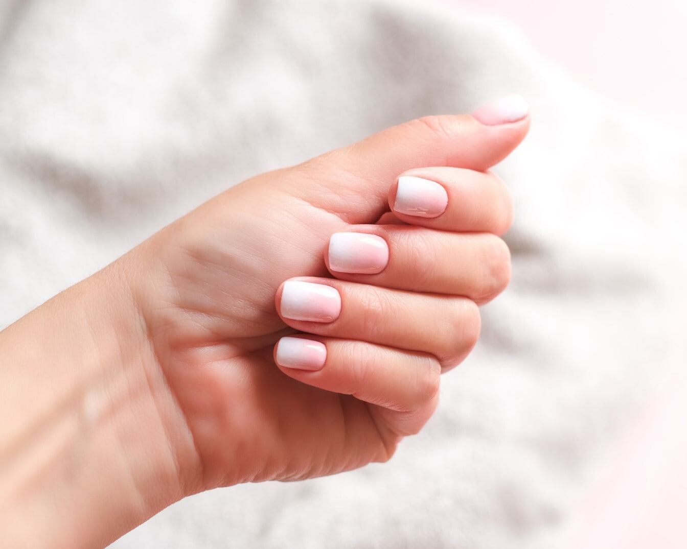 Delikatna dłoń z krótkimi, kwadratowymi paznokciami, pomalowanymi w stylu ombre od jasnoróżowego u nasady do białego na końcach, prezentowana na rozmytym, jasnym tle.