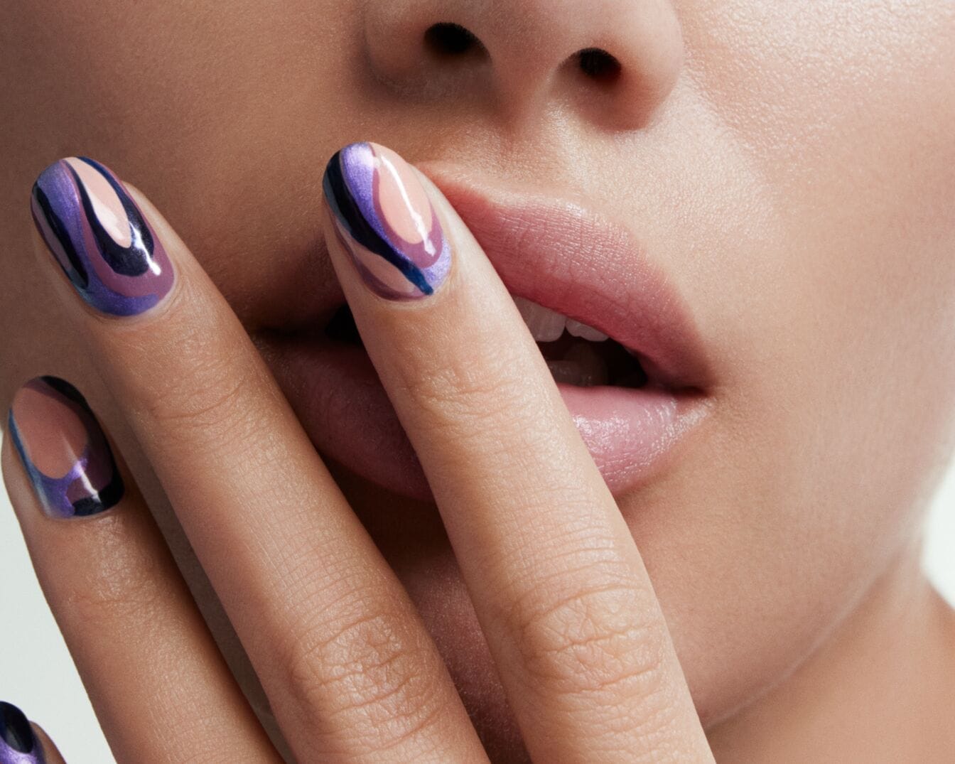 Zbliżenie na kobiecą dłoń z pomalowanymi paznokciami w abstrakcyjne wzory w odcieniach fioletu i beżu, dotykającą ust.