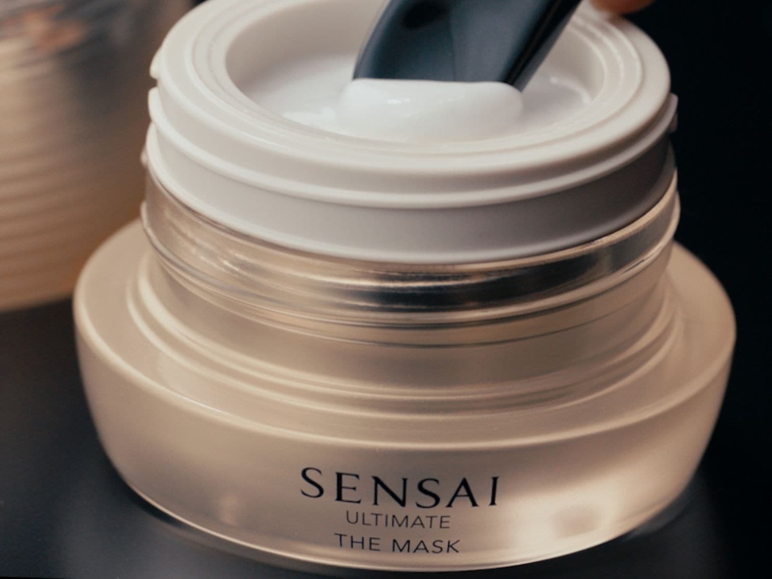 Detailní pohled na pleťovou masku Sensai Ultimate The Mask v průhledné skleněné dóze s bílým víčkem a špachtlí, která nabírá krémovou texturu.