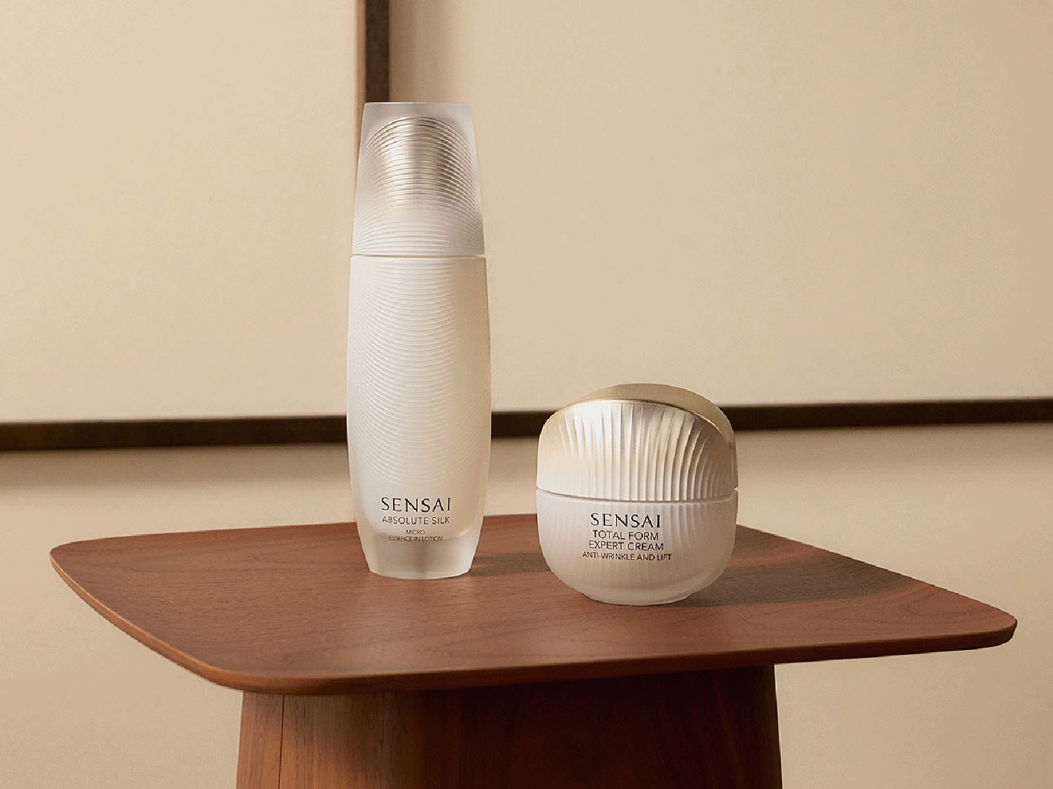 Dva produkty péče o pleť Sensai Absolute Silk a Total Form Expert Cream jsou elegantně umístěny na dřevěném stolku.